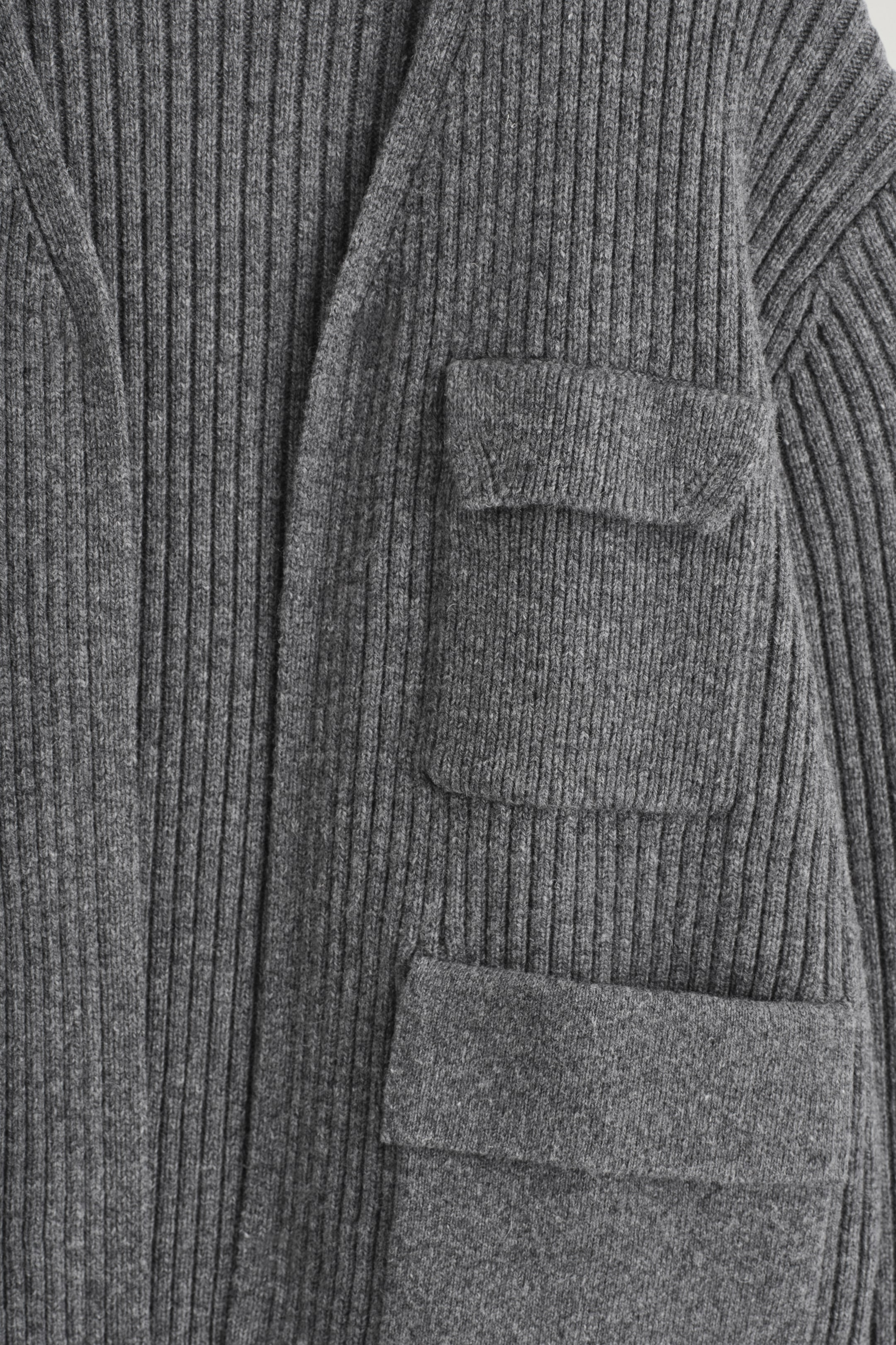 Visualizza immagine più grande: Un indumento grigio lavorato a maglia presenta una lavorazione a coste spesse, con due tasche a patta, una sopra l'altra, visibili sul davanti.