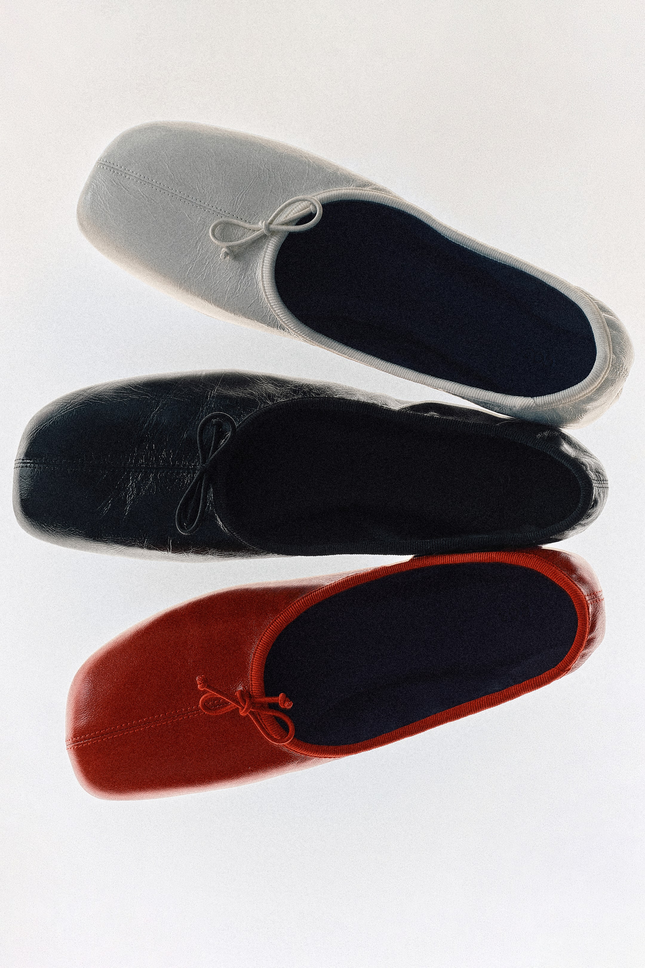 Agrandir l'image: Trois ballerines en cuir sont disposées verticalement, présentant une chaussure gris clair, noire et rouge vue de dessus. Chaque ballerine présente un bout carré encadré par des coutures apparentes, un bord en gros-grain et un nœud discret sur l’avant.