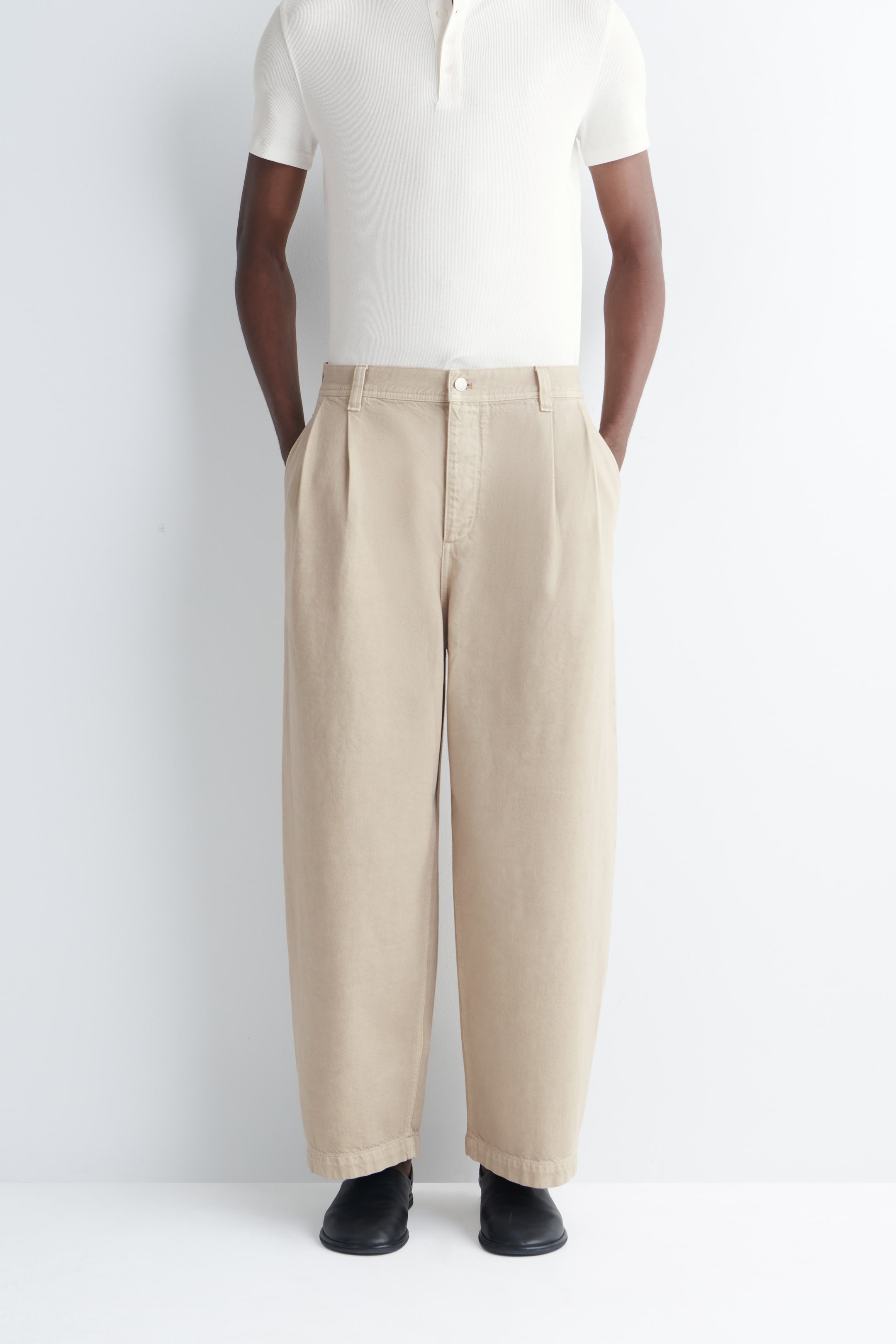 Grotere afbeelding bekijken: De model draagt beige broek met tonvormige pijpen. De broek heeft plooien aan de voorkant, riemlussen en een knoopsluiting, met pijpen die tot aan de enkel reiken. Het model staat met de handen in de zakken.