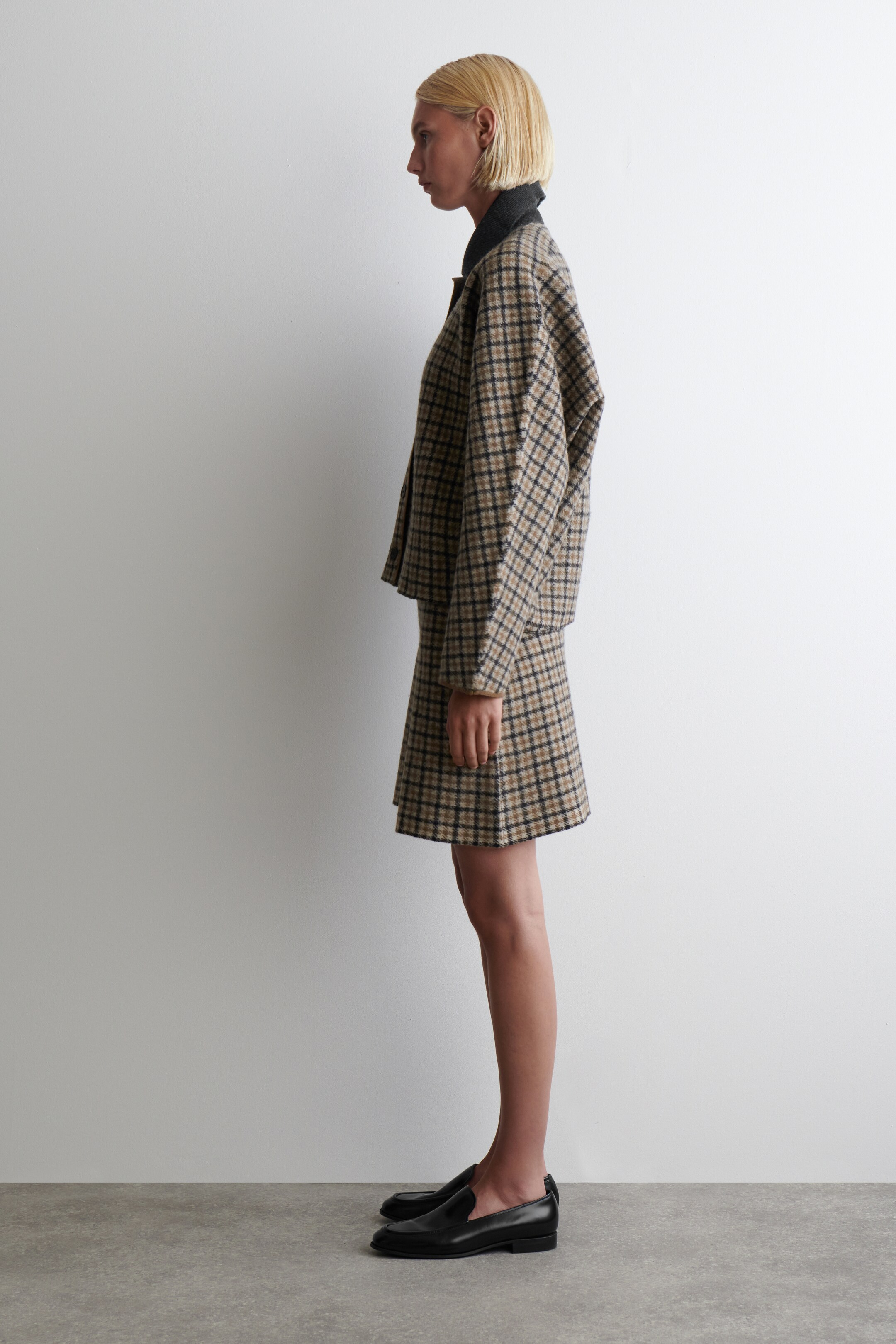 View larger image: A brown, beige, and black plaid jacket and matching mini skirt are being presented from a side angle. The jacket is featuring a relaxed, boxy fit with a dark grey underlayer visible at the collar, and the skirt is showing an A-line silhouette.