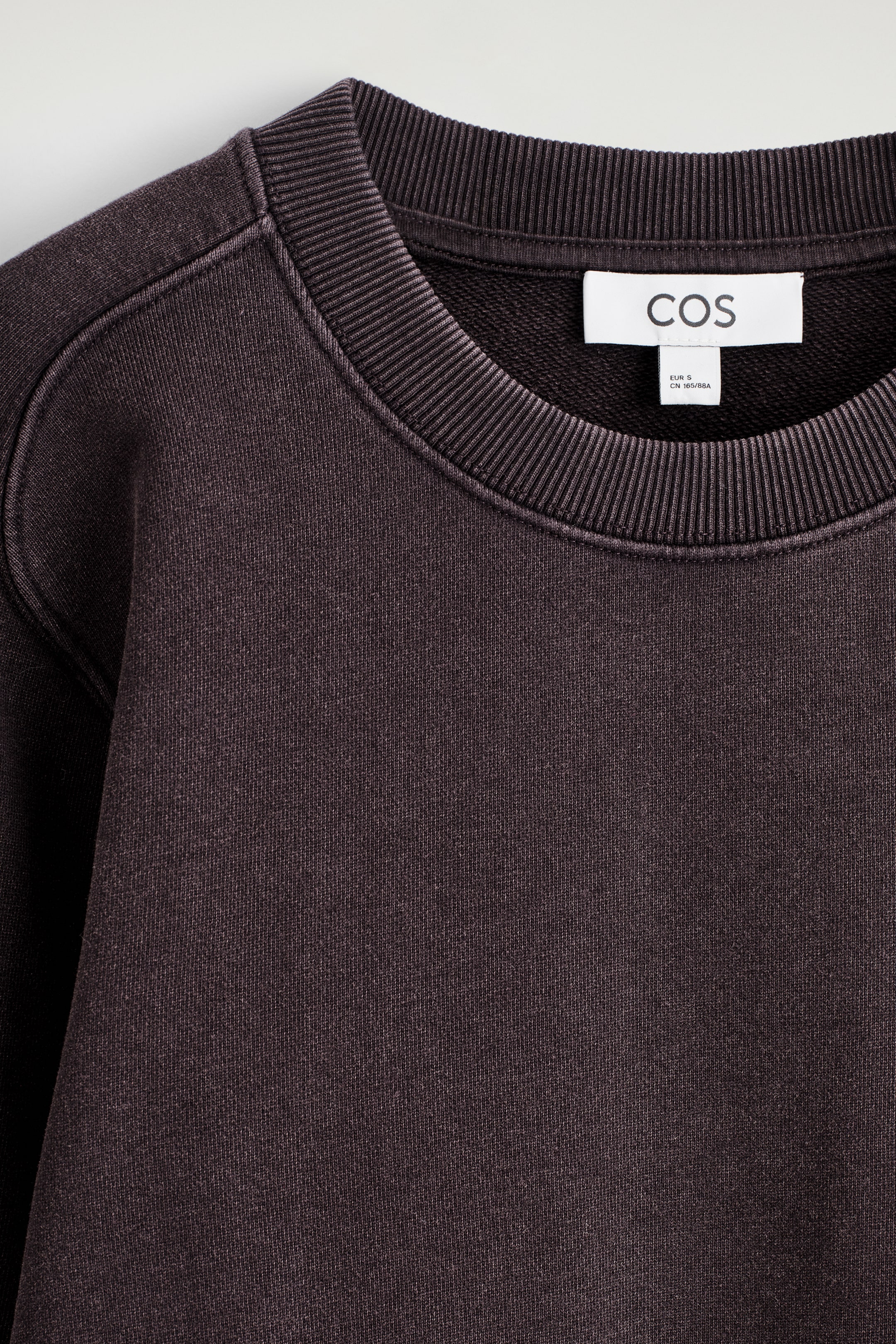Agrandir l'image: Un sweat-shirt en coton gris foncé est présenté de face, avec un col rond côtelé. Une étiquette de marque COS blanche est visible à l'arrière du cou, et le tissu présente une texture douce, légèrement délavée.
