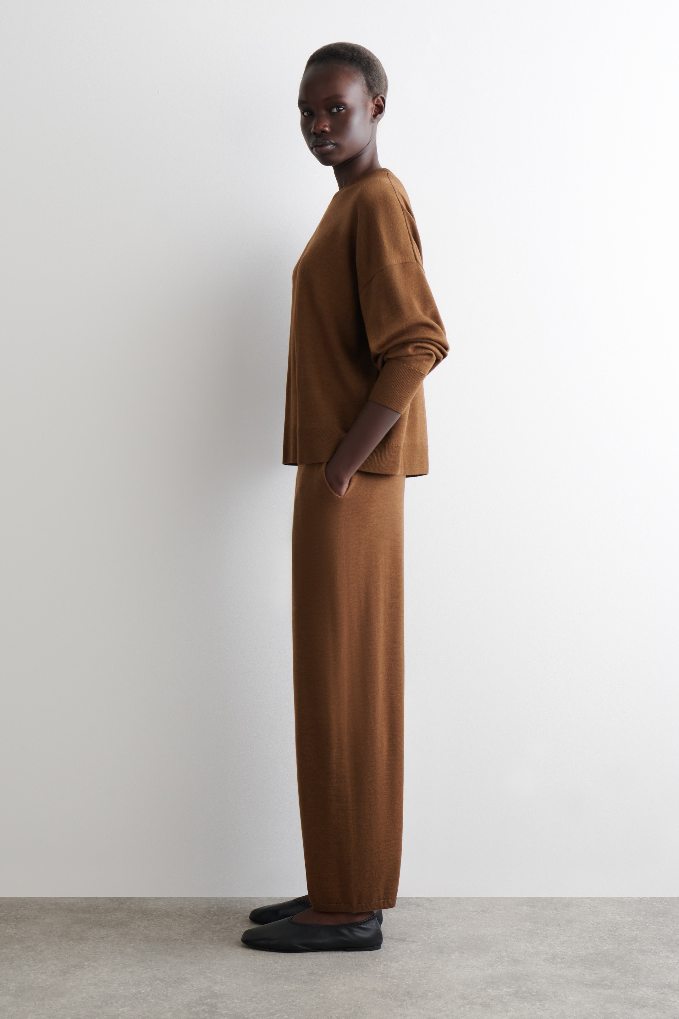 Ingrandisci l'immagine: Un completo marrone a maglia in due pezzi viene presentato di profilo, caratterizzato da un top a V con spalle scese e polsini a coste, abbinato a pantaloni a gamba dritta coordinati. Con l'outfit vengono indossate delle ballerine nere.