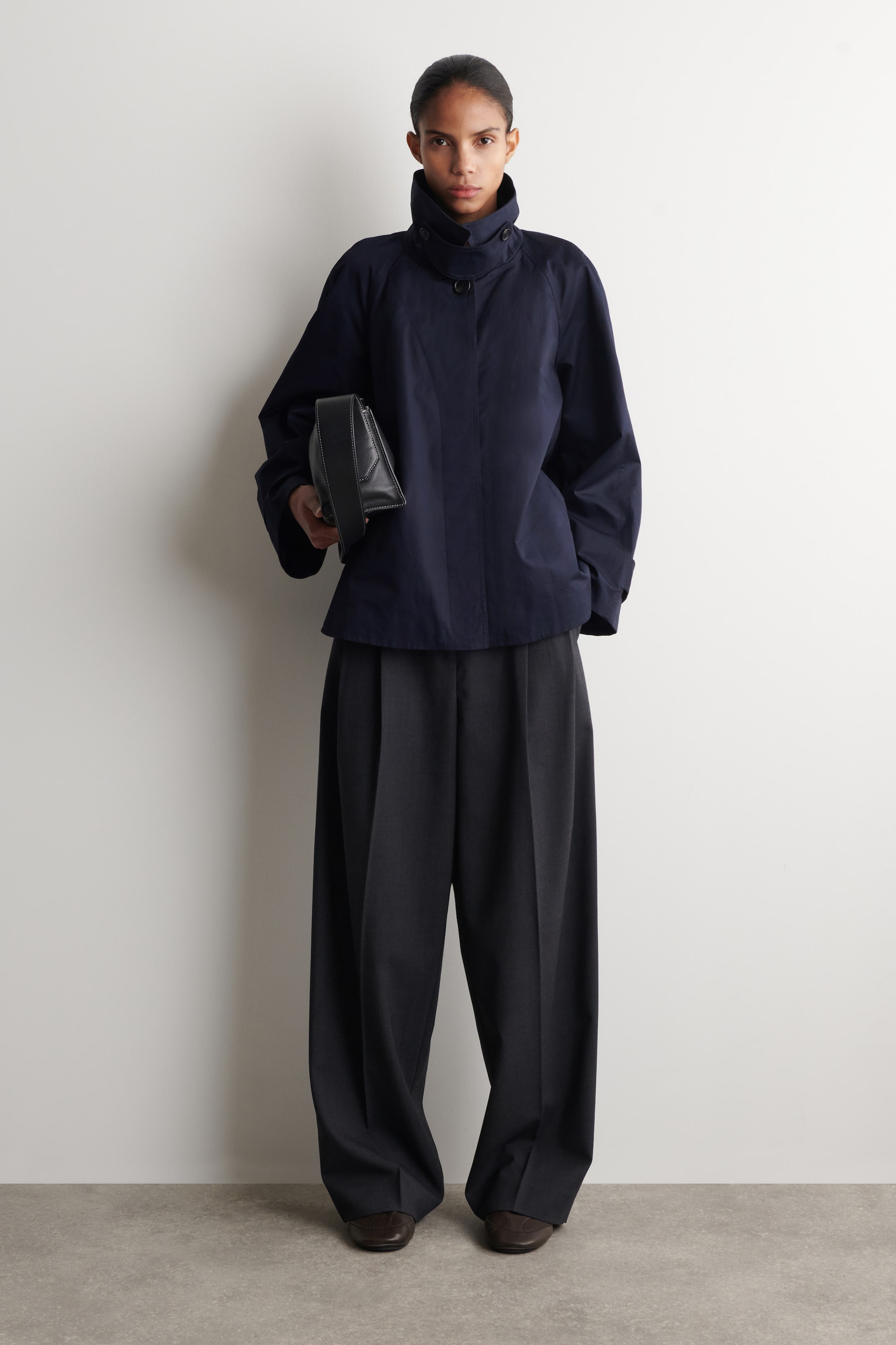 Näytä suurempi kuva: Tummansininen takki, jossa on korkea pystykaulus ja näkyvät napit sekä leveät raglanhihat. Takin kanssa on yhdistetty tummanharmaat, leveälahkeiset laskostetut housut, jotka laskeutuvat tummanruskeiden teksturoitujen kenkien päälle.