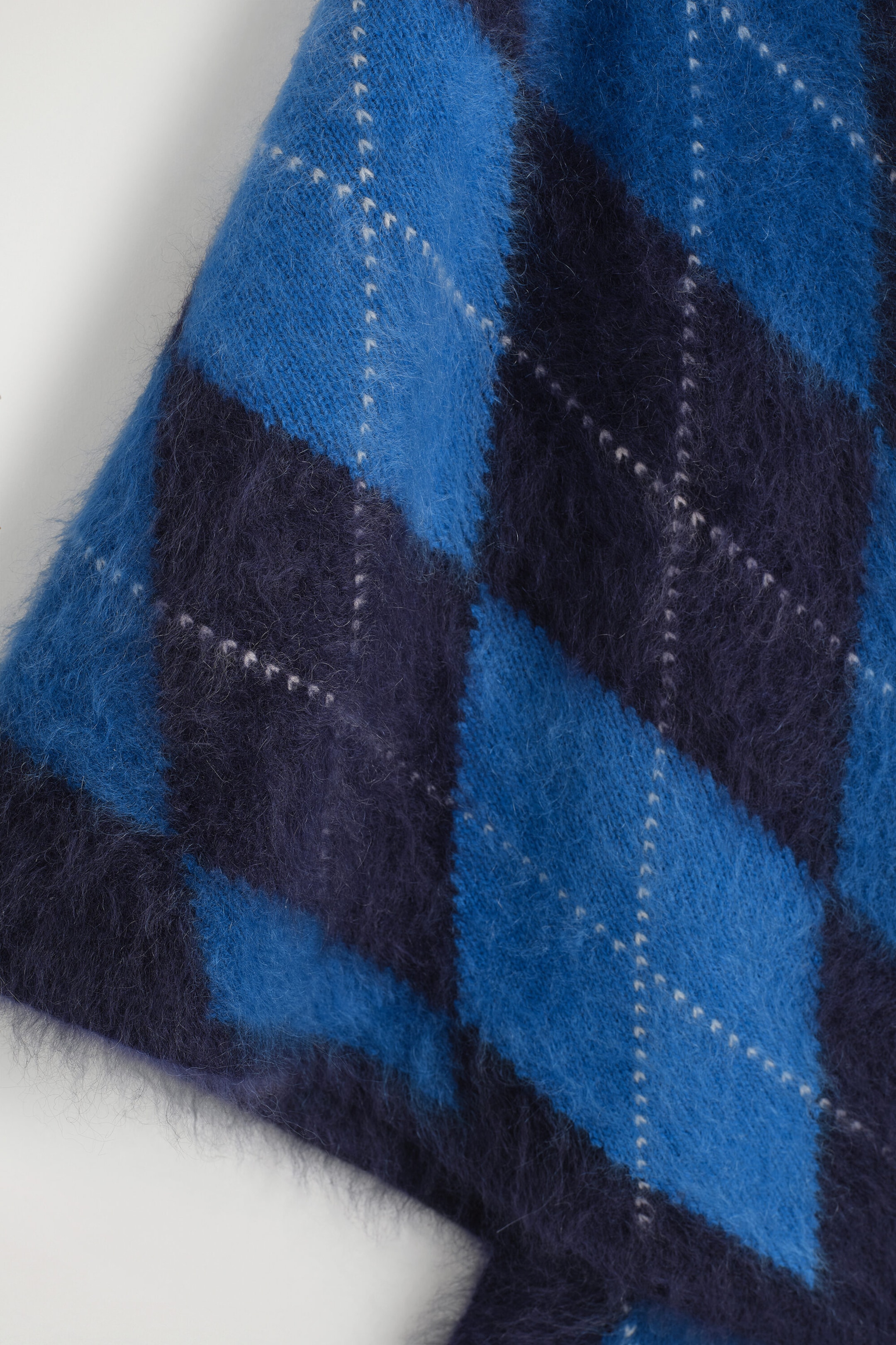 The Argyle Brushed-Cashmere Triangle Scarf is showing its soft, brushed texture and classic argyle pattern in a close-up view. The design features large diamonds in bright blue and dark navy, intersected by thin, dotted white lines. The Argyle Brushed-Cashmere Triangle Scarf is showing its soft, brushed texture and classic argyle pattern in a close-up view. The design features large diamonds in bright blue and dark navy, intersected by thin, dotted white lines.