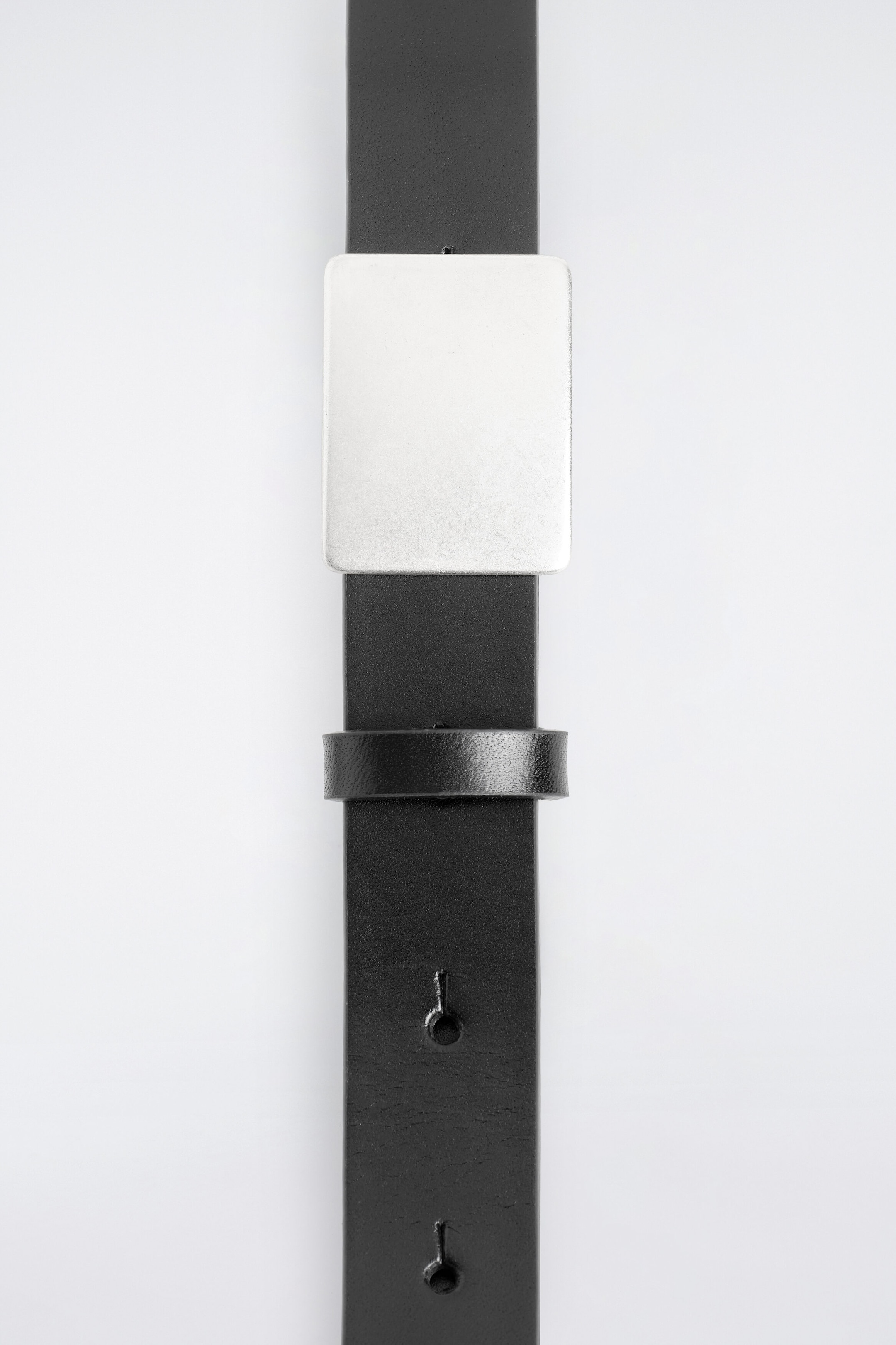 A leather belt with a metal buckle is shown extending vertically in a front-facing view, showcasing a smooth black leather strap and a square, silver-toned metal buckle with a matte finish. The strap features a matching leather keeper loop and keyhole-style adjustment holes below the buckle.