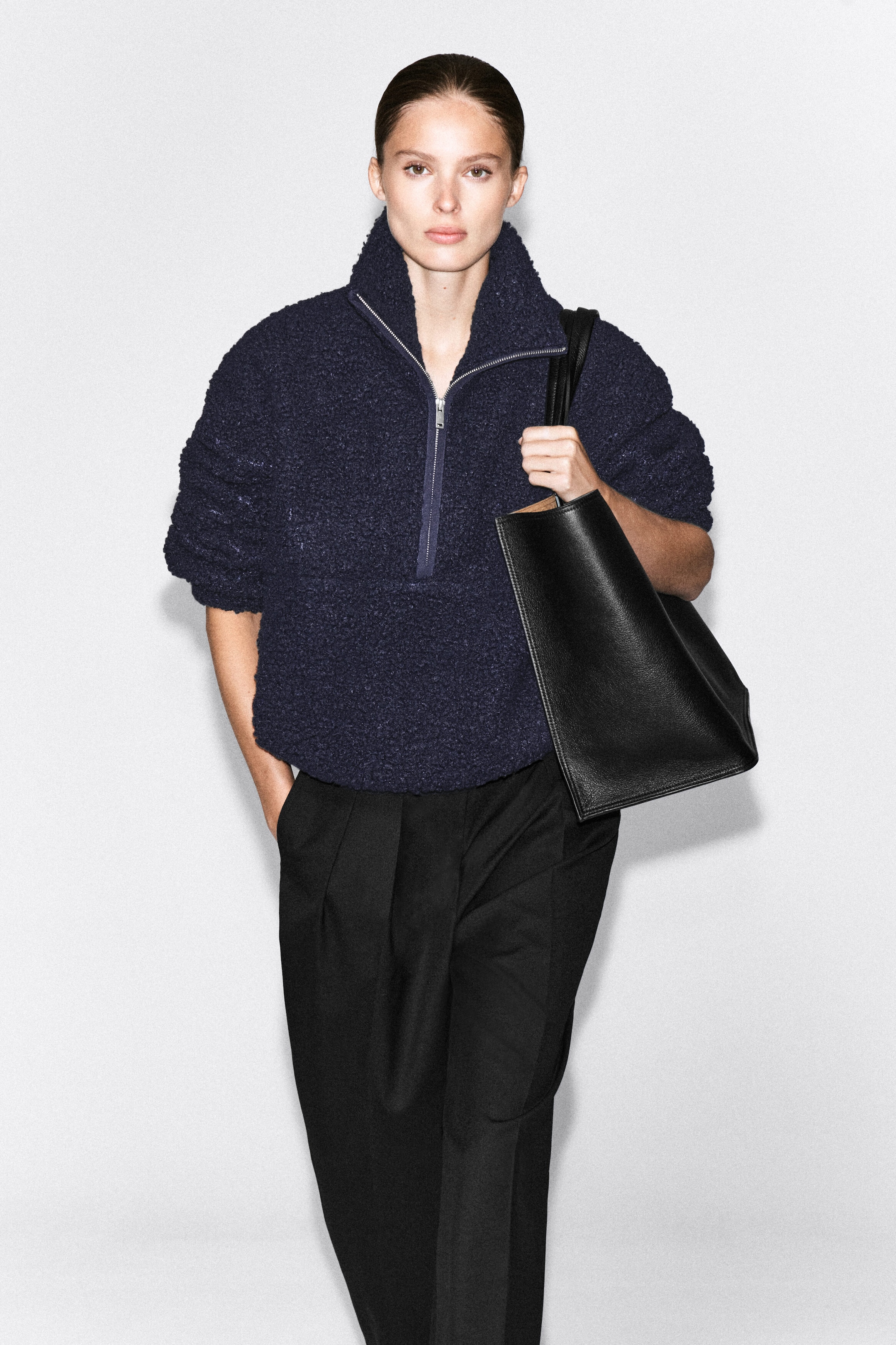 The model is wearing a dark navy blue teddy fleece half-zip jumper, featuring a relaxed fit and a high funnel neck with a silver zipper. The jumper is paired with black trousers, and the model is carrying a black leather tote bag over one shoulder. The model is wearing a dark navy blue teddy fleece half-zip jumper, featuring a relaxed fit and a high funnel neck with a silver zipper. The jumper is paired with black trousers, and the model is carrying a black leather tote bag over one shoulder.