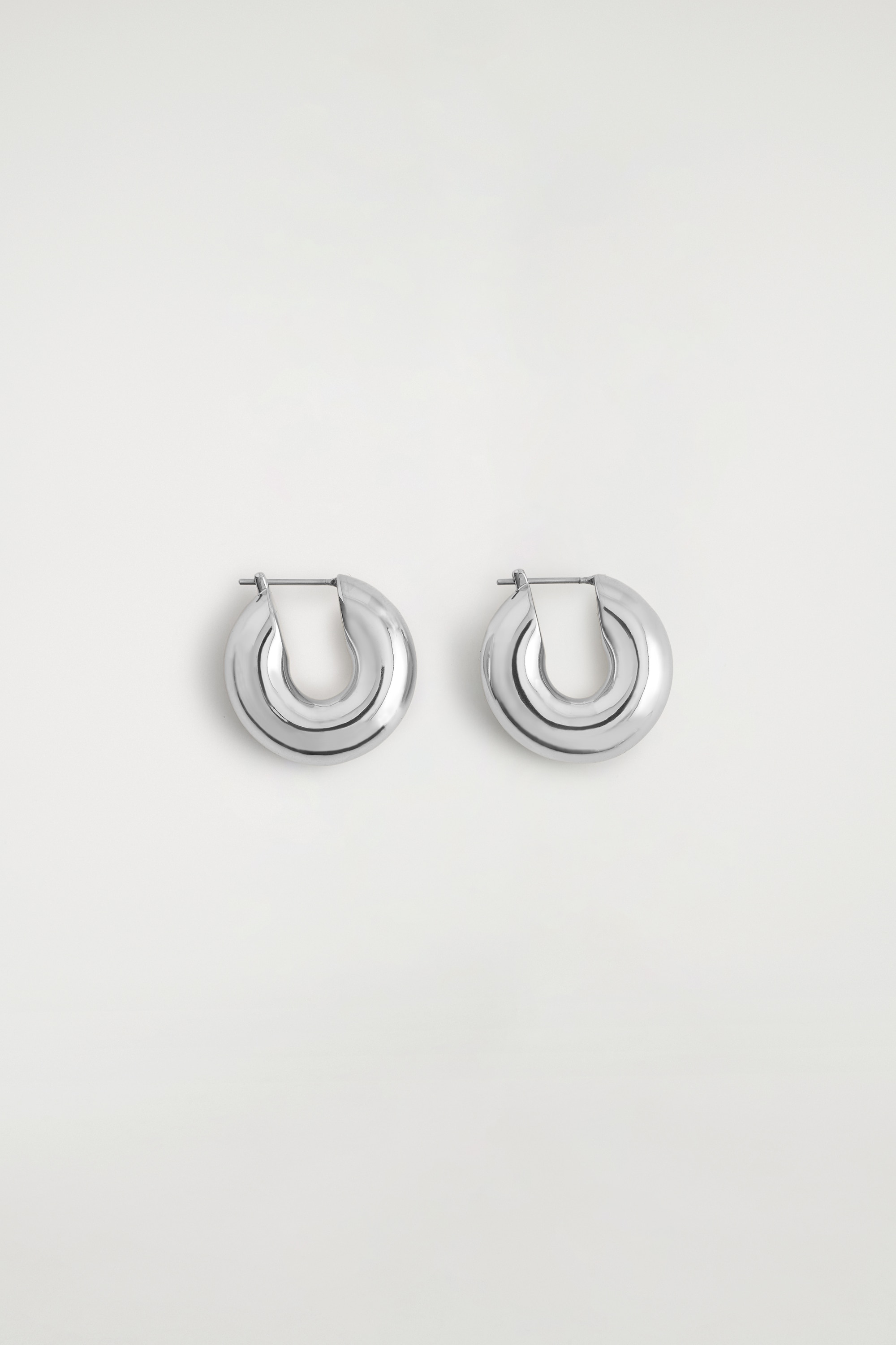 Brincos pequenos de aro, em metal prateado polido, estÃ£o a ser usados nos lÃ³bulos das orelhas da modelo. Os aros pequenos e espessos sÃ£o combinados com o cabelo apanhado para trÃ¡s da modelo e uma camisola transparente azul-escura.