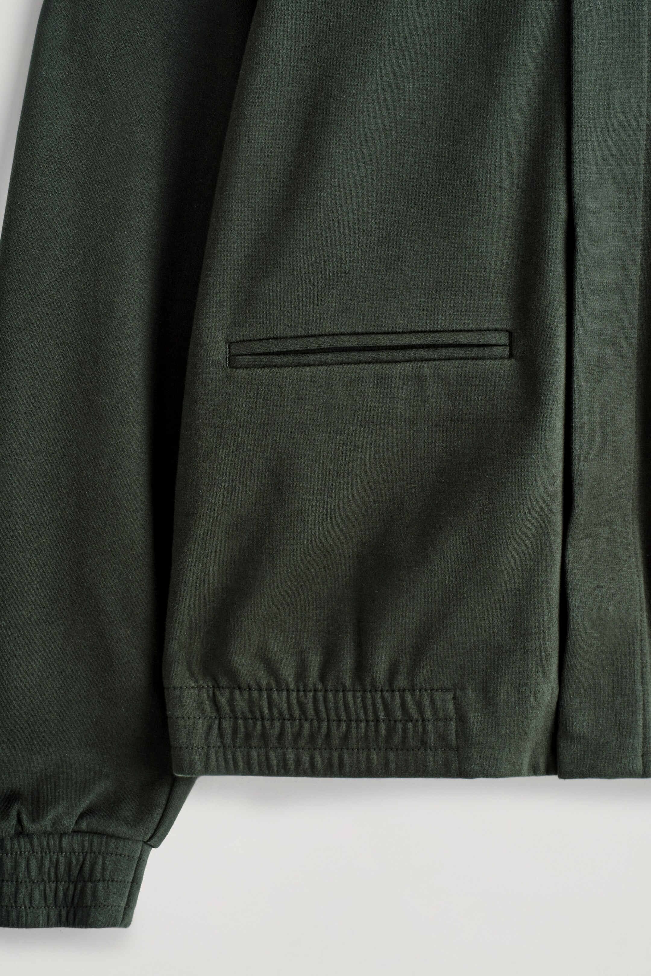 Agrandir l'image: Une veste en jersey vert foncé présente une poche passepoilée horizontale sur le devant inférieur. Des détails élastiqués sont visibles sur l'ourlet du bas et le poignet de la manche.