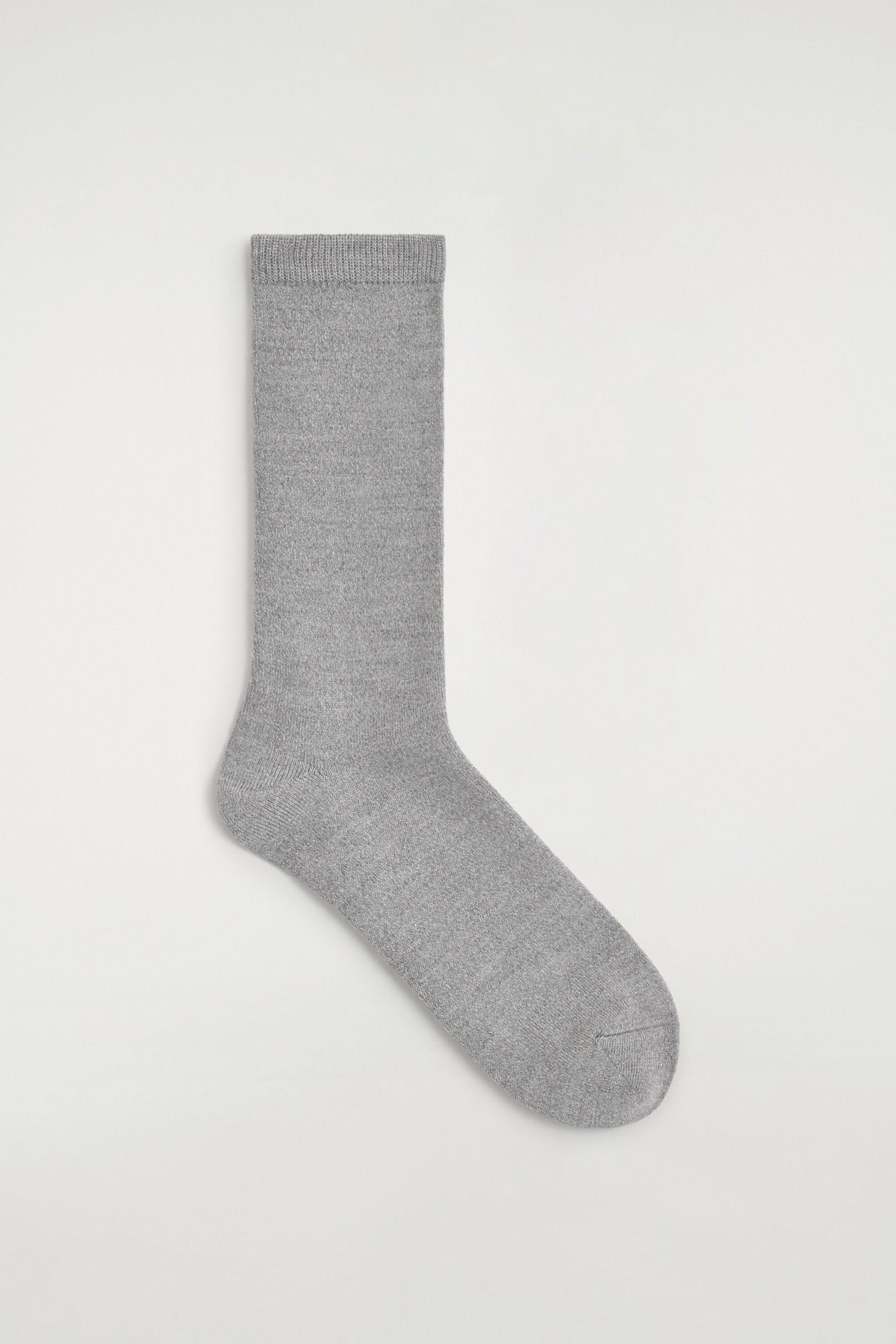Une fine chaussette en laine pailletée est posée à plat, vue de profil, mettant en valeur sa maille fine gris clair avec un léger reflet métallisé. La chaussette présente une bordure côtelée en haut, un talon profilé et un bout arrondi.