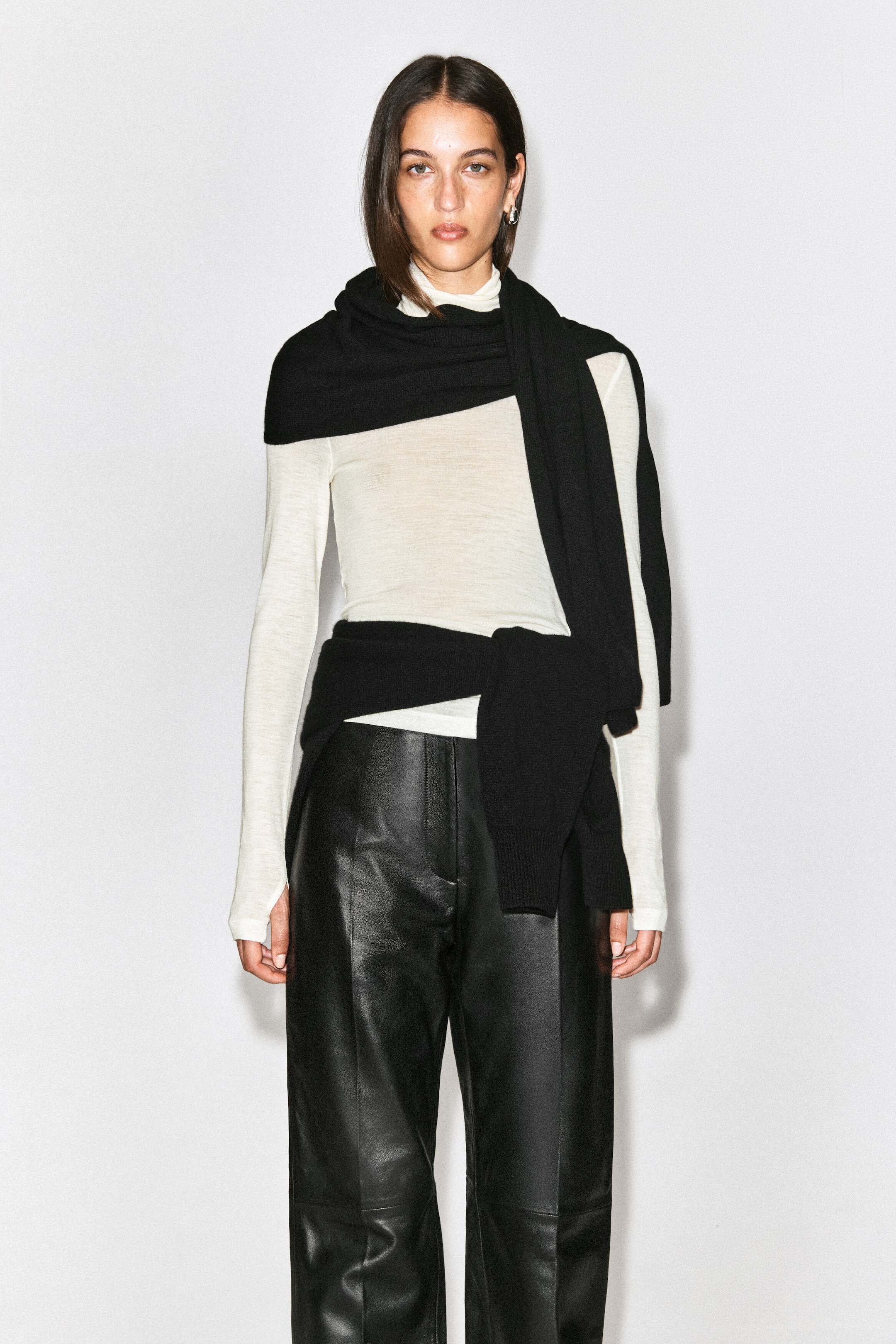 A black cashmere scarf is being worn wrapped around the neck and draped over the shoulders of the model. The scarf is layered over a cream-coloured, long-sleeve top and styled with black leather trousers.