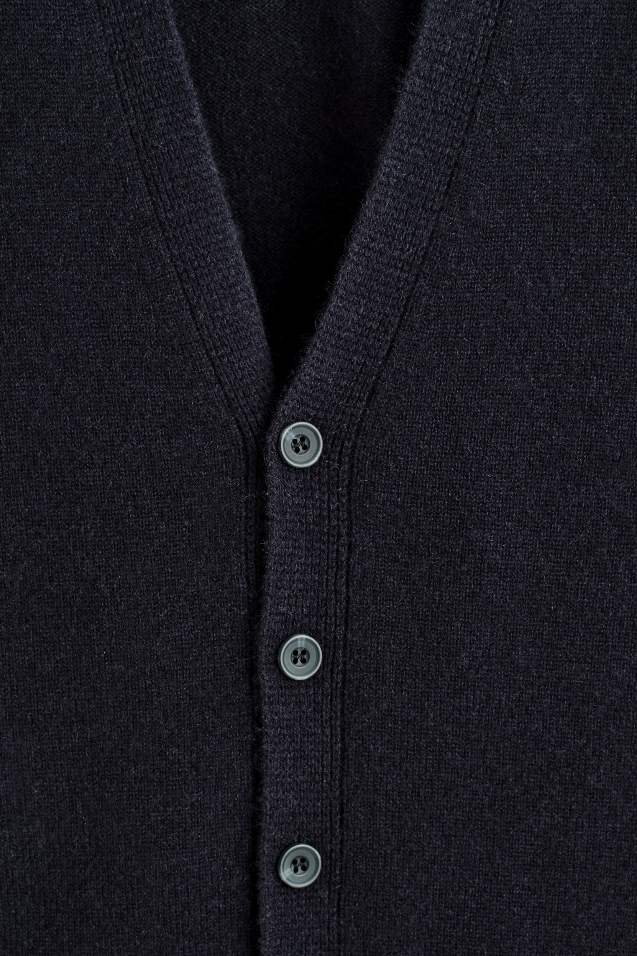 Größeres Bild ansehen: Ein marineblauer Kaschmir-Cardigan wird von vorne gezeigt, mit einem V-Ausschnitt und einer zentralen Knopfleiste. Mehrere runde, graue Knöpfe sind entlang der Leiste sichtbar, die sich gegen die feine Strickstruktur des Stoffes abheben.