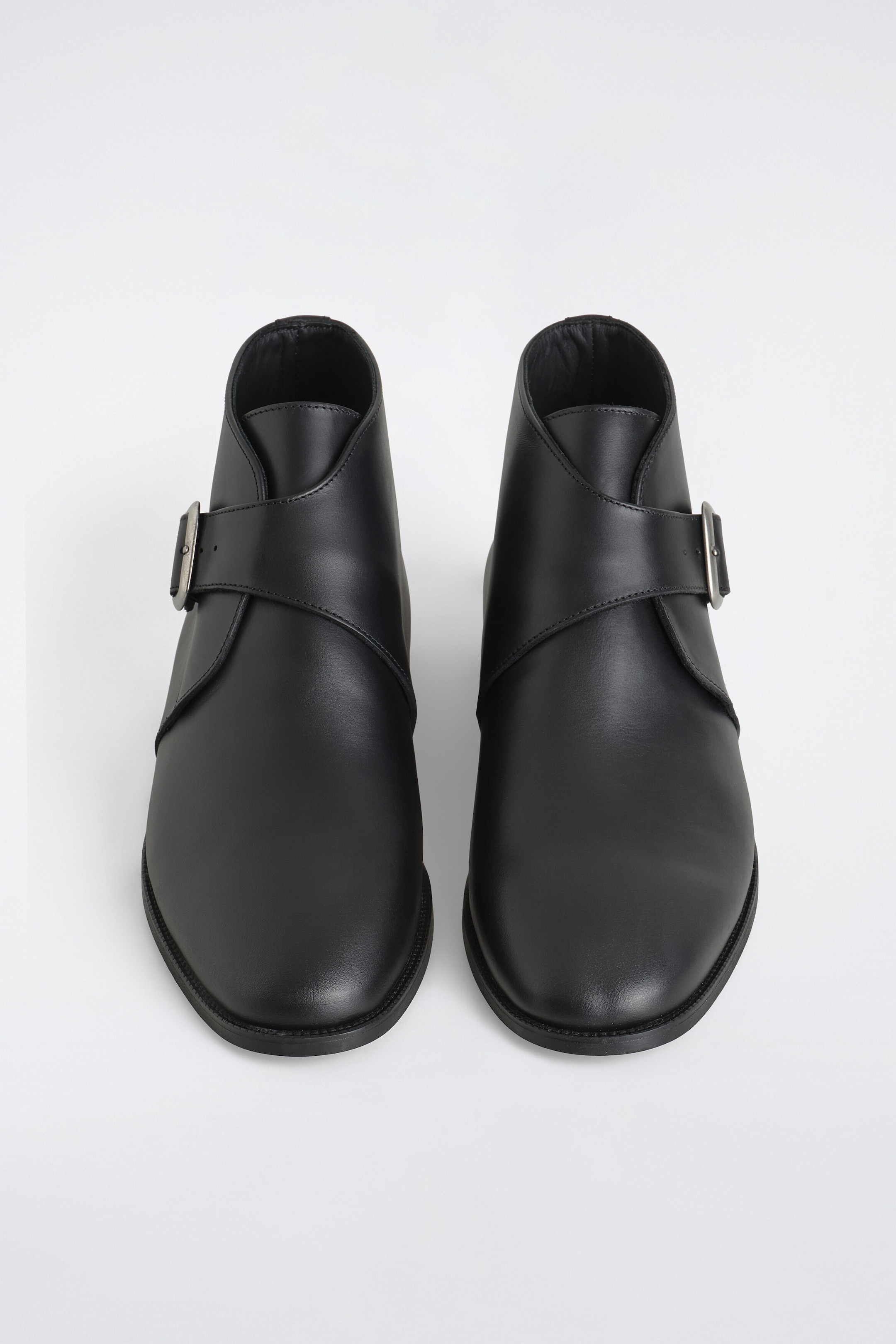 Ein Paar schwarze Leder-Monk-Boots ist frontal ausgerichtet und verfügt über ein glattes Lederoberteil sowie abgerundete Zehen. Die knöchelhohen Stiefel zeigen einen breiten, überkreuzten Riemen, der sich um die Vorderseite des Knöchels legt und an der Seite mit einer silberfarbenen Metallschnalle befestigt ist.