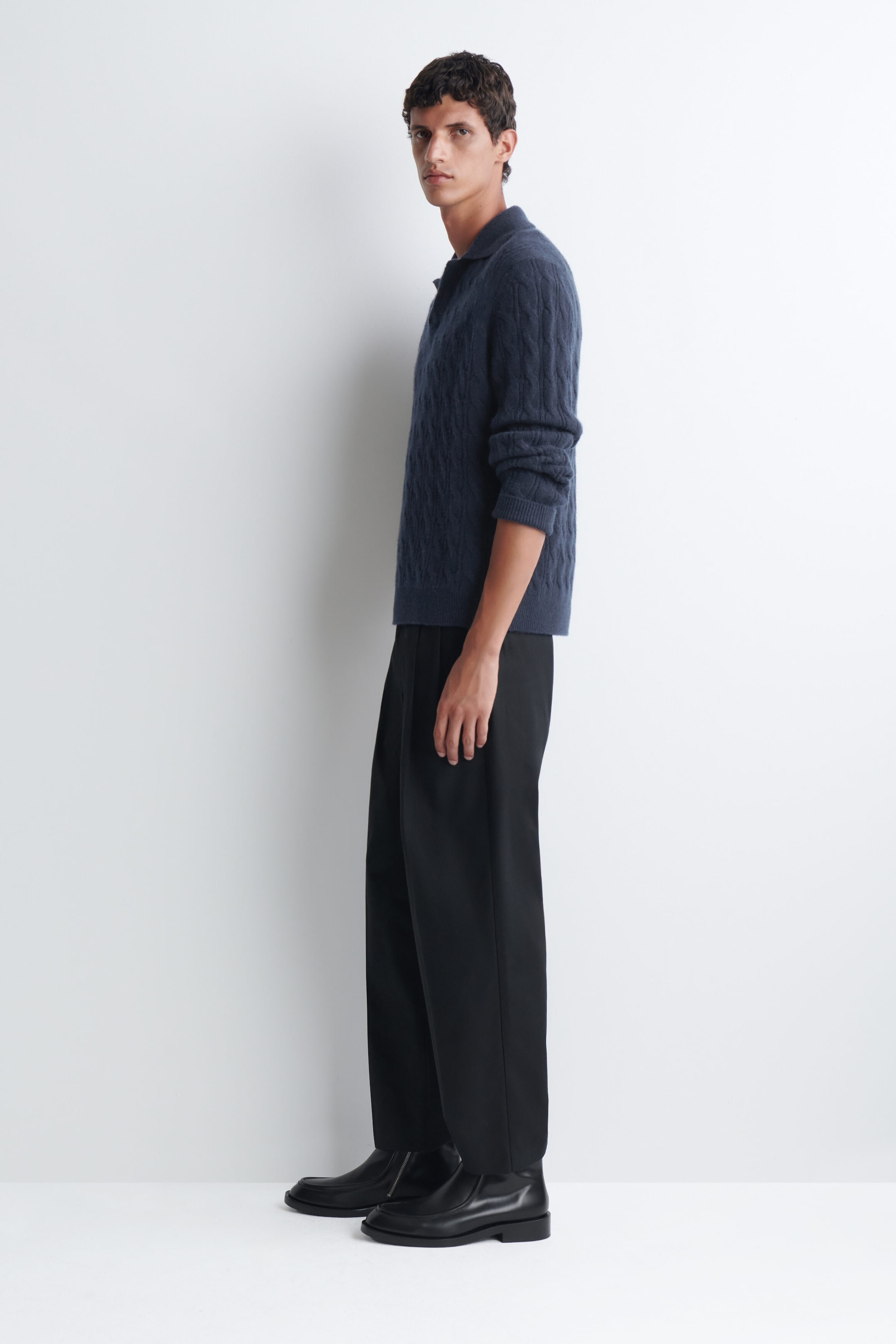 A cable-knit cashmere polo sweater in dark blue is being worn by the model, featuring a classic collar, long sleeves, and a textured knit pattern. The sweater is paired with black wide-leg trousers and black leather boots.