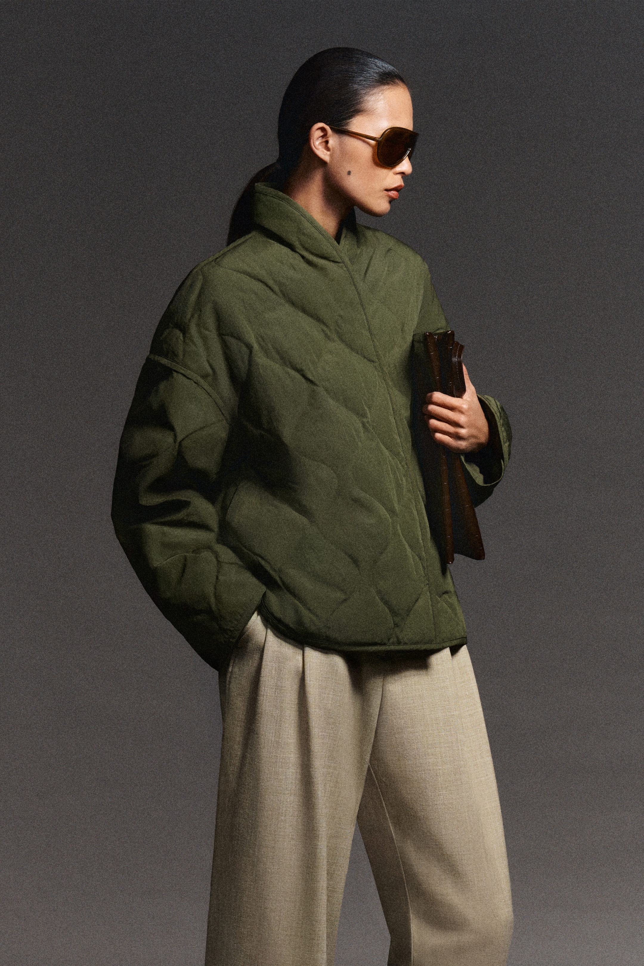 The olive green shawl-collar quilted jacket is showcased from a three-quarter front angle, highlighting its diamond-quilted design and relaxed fit. It features a high shawl collar, a curved hem, and long sleeves, styled with light beige wide-leg trousers.