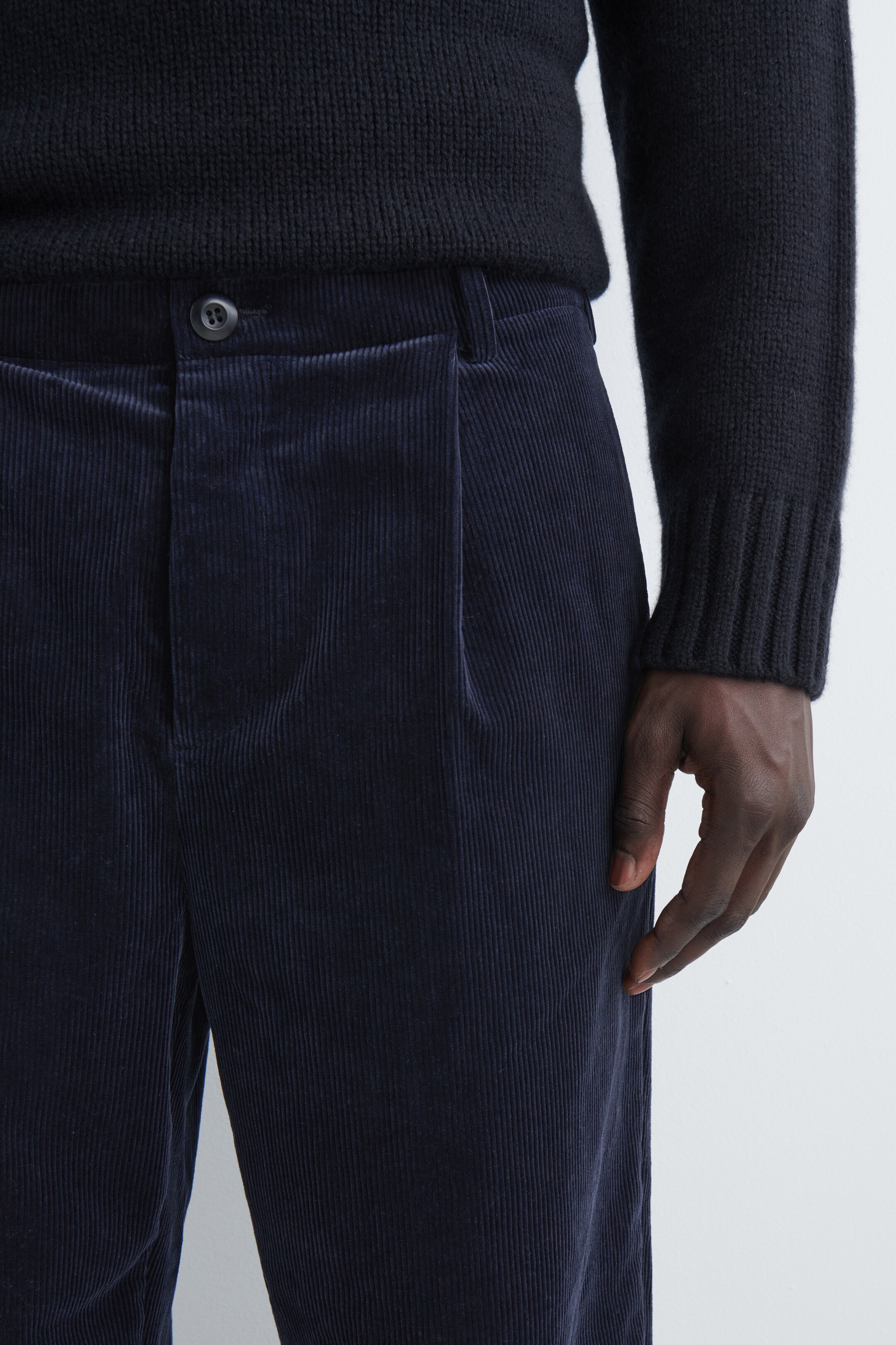 Agrandir l'image: Un pantalon en velours côtelé bleu foncé est présenté sous un angle incliné, avec un seul pli à l'avant et une fermeture à bouton à la taille. Une main repose dans une poche latérale, et un pull en tricot foncé est superposé au-dessus de la ceinture.