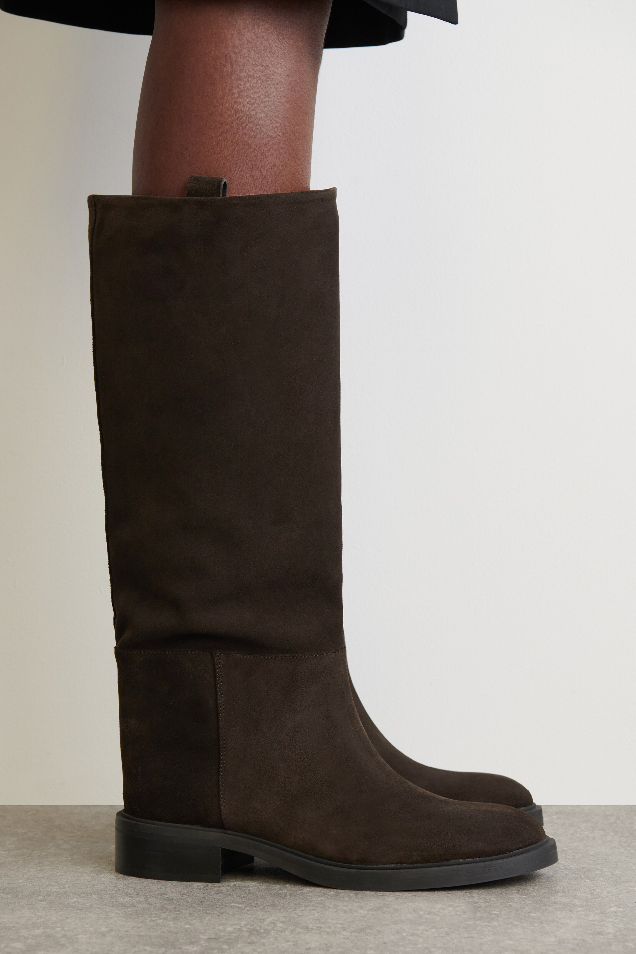 Größeres Bild ansehen: Ein dunkelbrauner Wildlederstiefel wird getragen und in einer Seitenansicht präsentiert. Der Stiefel verfügt über einen glatten, unstrukturierten Schaft, der bis zum Knie reicht, eine abgerundete Spitze, einen niedrigen schwarzen Blockabsatz mit flacher Sohle und eine kleine Schlaufe, die am inneren oberen Rand sichtbar ist.