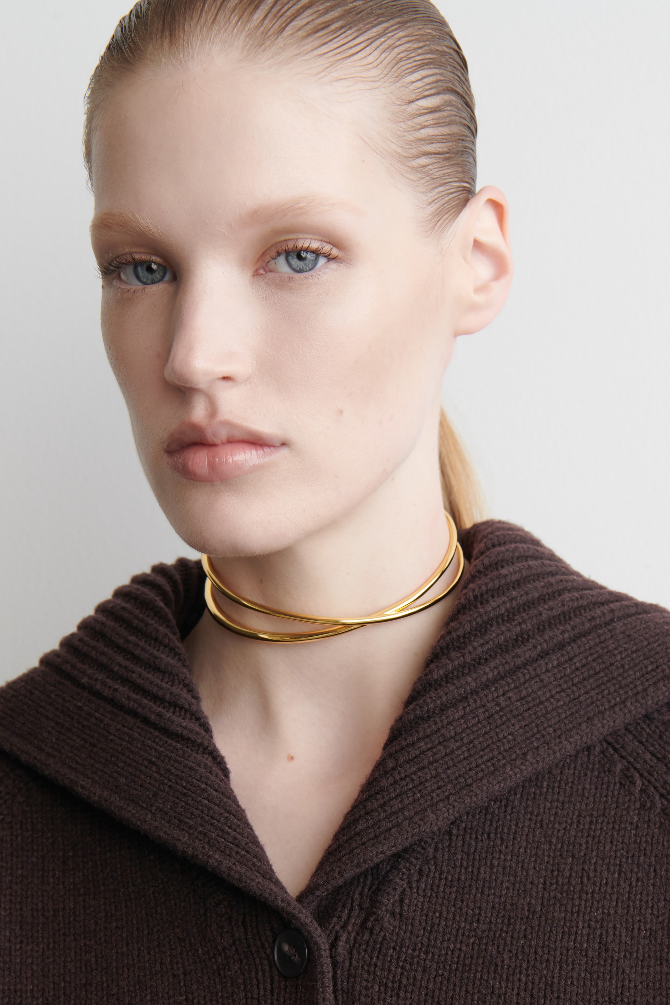Visualizza immagine più grande: Un collare dorato presenta due bande sottili che si incrociano l'una sull'altra, creando un design intrecciato intorno alla base del collo e poggiando sopra un colletto marrone scuro lavorato a maglia.