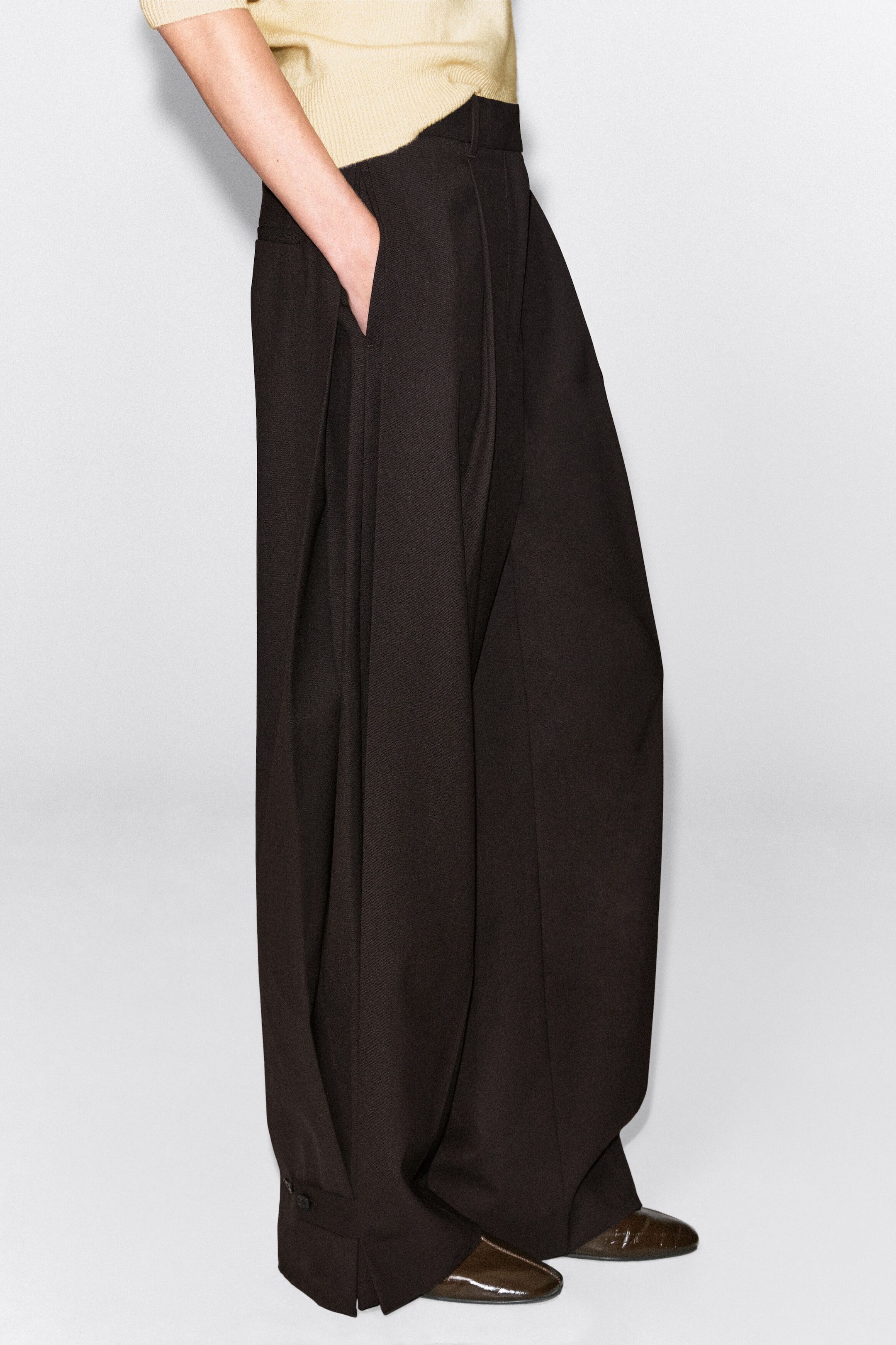 Dunkelbraune, weit geschnittene Hose mit hoher Taille und Bügelfalten, getragen von dem Model, das mit einer Hand in der Seitentasche steht. Die Hose zeichnet sich durch eine drapierte Silhouette mit Knopfleisten-Details an den geschlitzten Säumen aus und wird mit einem in den Bund gesteckten hellgelben Stricktop kombiniert. Dunkelbraune, weit geschnittene Hose mit hoher Taille und Bügelfalten, getragen von dem Model, das mit einer Hand in der Seitentasche steht. Die Hose zeichnet sich durch eine drapierte Silhouette mit Knopfleisten-Details an den geschlitzten Säumen aus und wird mit einem in den Bund gesteckten hellgelben Stricktop kombiniert.