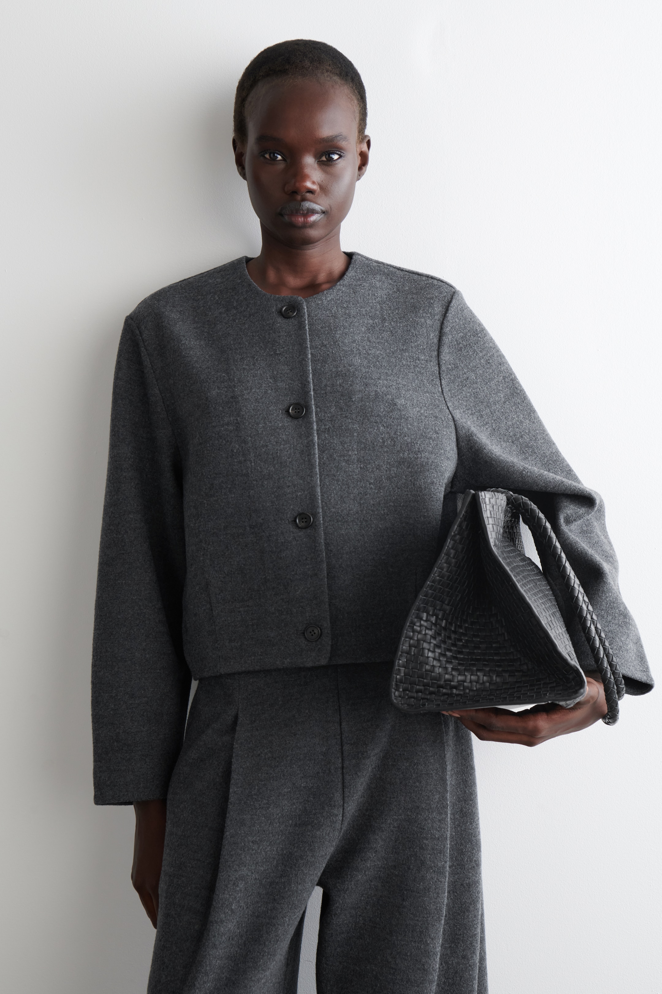 Agrandir l'image: Une veste courte sans col de couleur gris foncé avec une fermeture à trois boutons est présentée avec un pantalon plissé assorti de la même couleur, tandis qu'un sac à main noir tressé avec une poignée enroulée est tenu sur le côté.