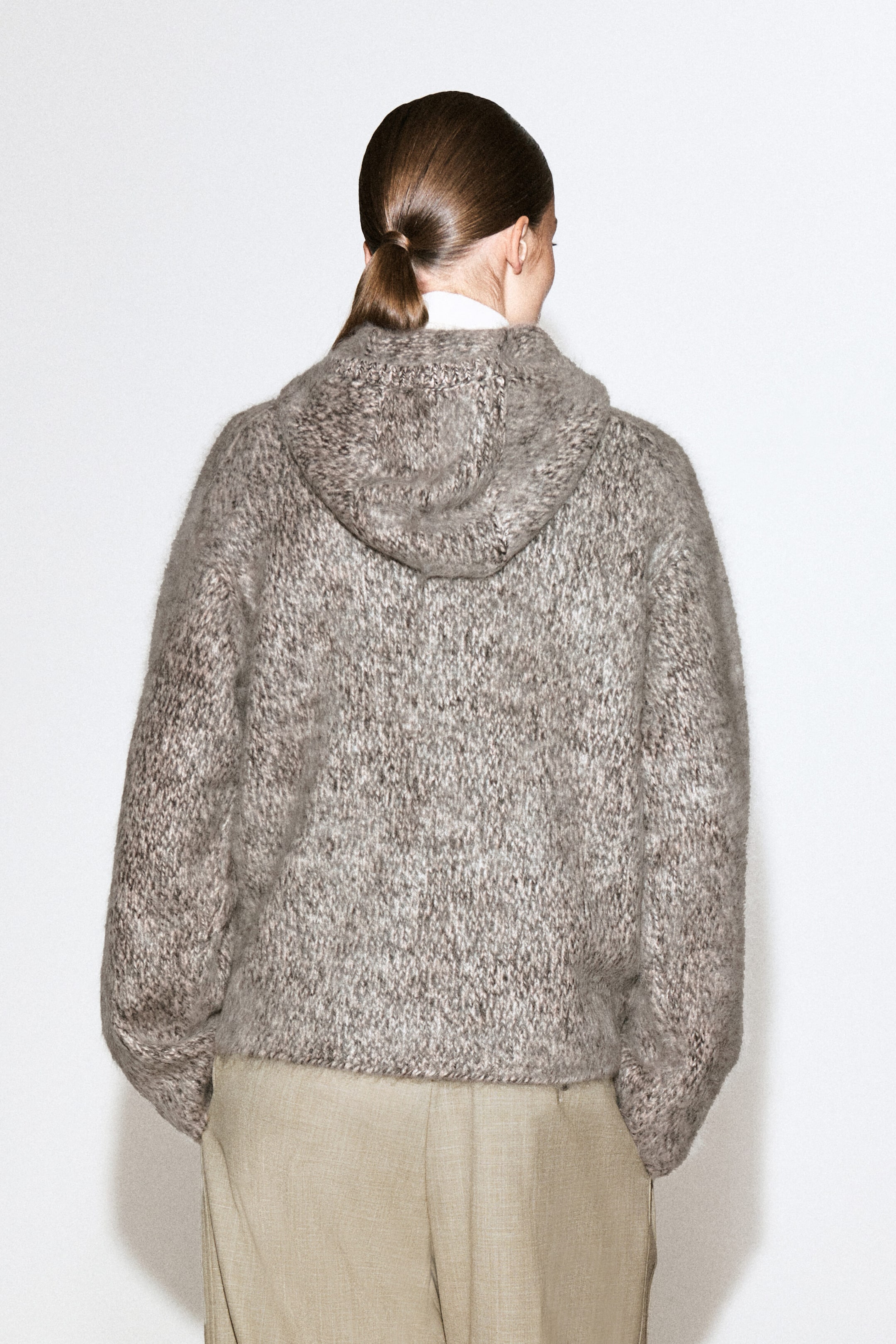 A chunky brushed-cashmere hoodie in a textured melange-brown knit is worn by the model, who is facing away to showcase the back of the garment and its soft hood. The hoodie is layered over a high white collar and styled with light beige trousers. A chunky brushed-cashmere hoodie in a textured melange-brown knit is worn by the model, who is facing away to showcase the back of the garment and its soft hood. The hoodie is layered over a high white collar and styled with light beige trousers.