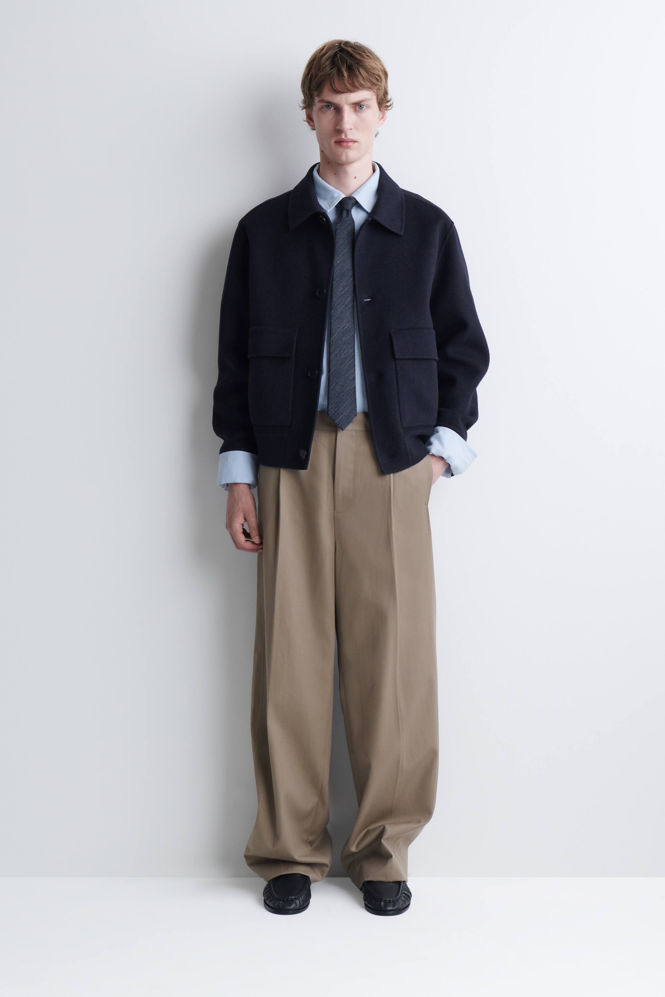 Agrandir l'image: Une cravate en soie-coton effet denim, dans une teinte gris-bleu foncé texturée, est portée par le mannequin. La cravate est stylisée sous le col d’une chemise bleu clair boutonnée et portée sous une veste foncée ouverte.