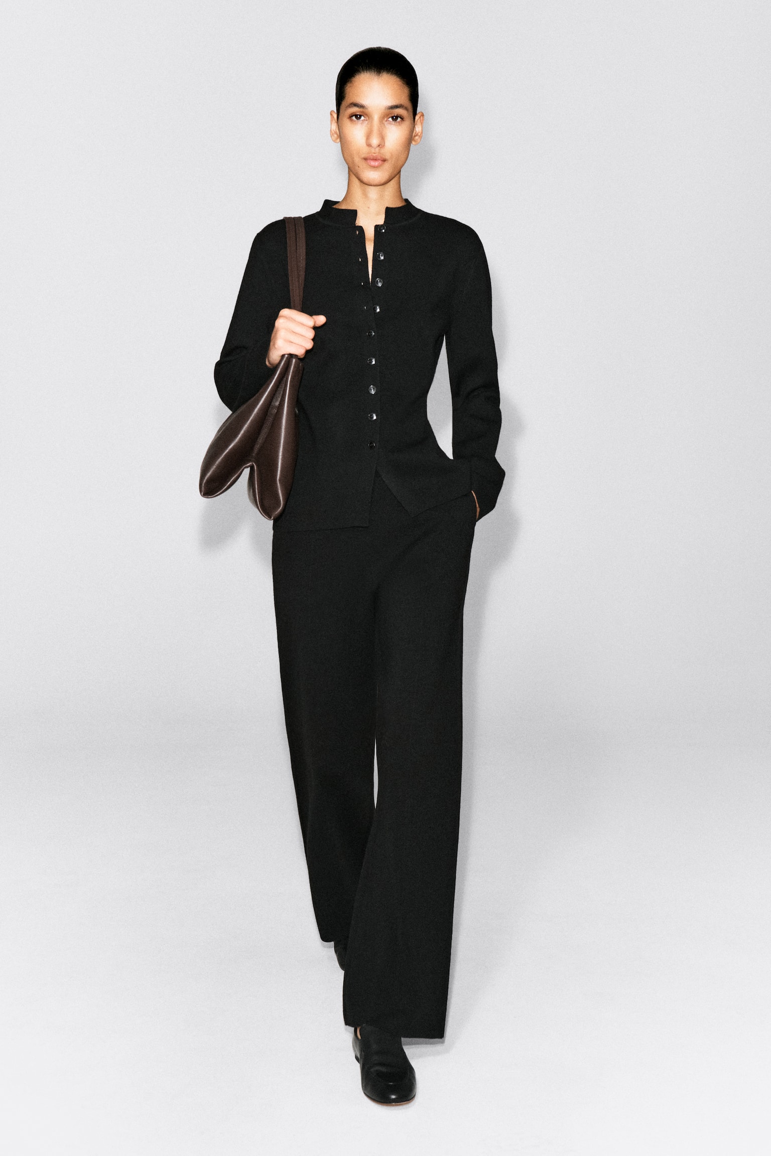 Le pantalon droit en mélange de laine mérinos est présenté de face, avec une silhouette décontractée à coupe droite en noir. Le pantalon tombe sur toute sa longueur sur des chaussures noires et est associé à un cardigan boutonné assorti, superposé à la taille. - 1