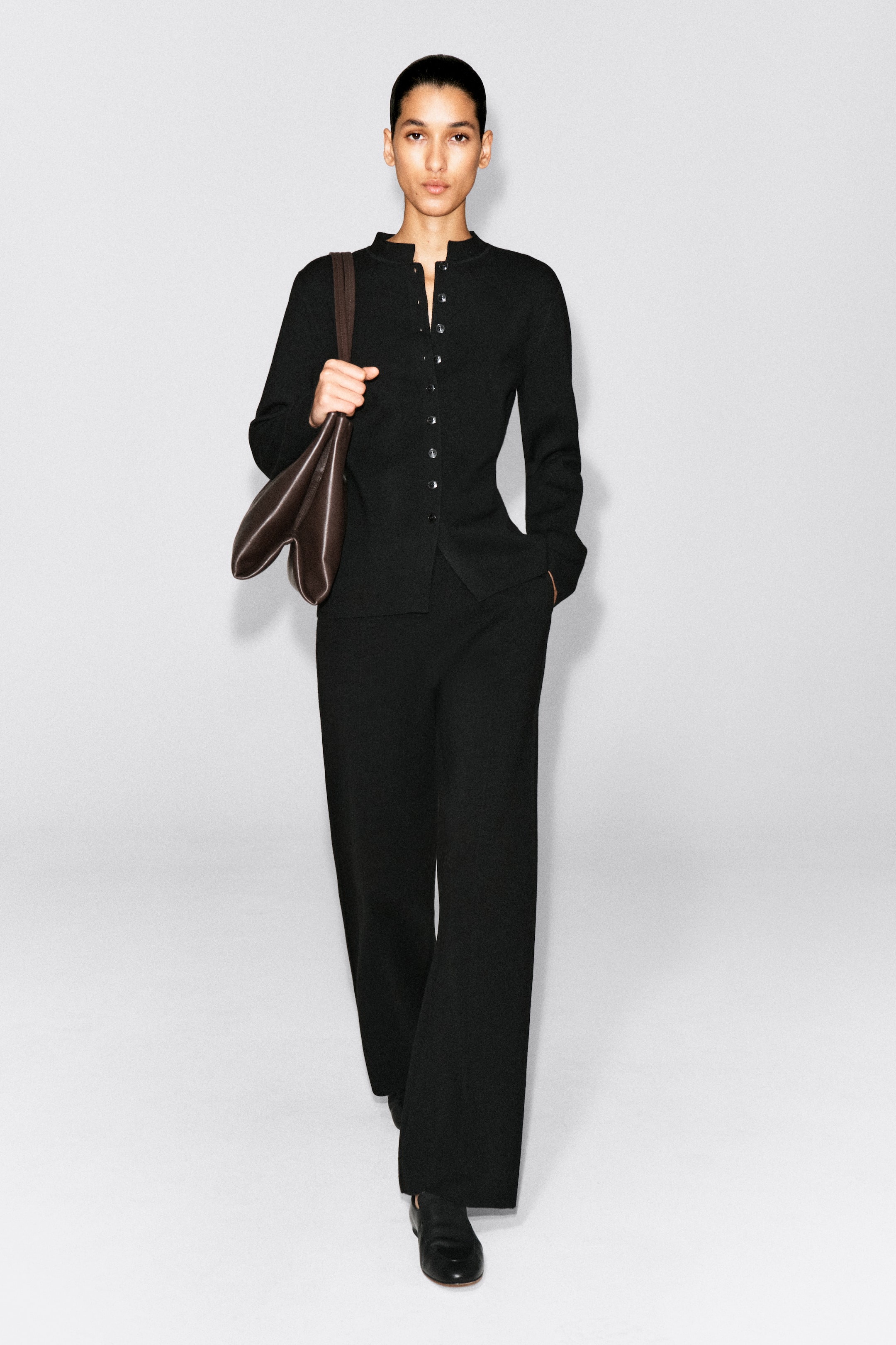 Visualizza l'immagine ingrandita: I pantaloni dritti in misto lana merino sono mostrati frontalmente, con una silhouette rilassata a taglio dritto in nero. I pantaloni arrivano a tutta lunghezza sopra scarpe nere e sono abbinati a un cardigan con bottoni coordinato, sovrapposto alla cintura.
