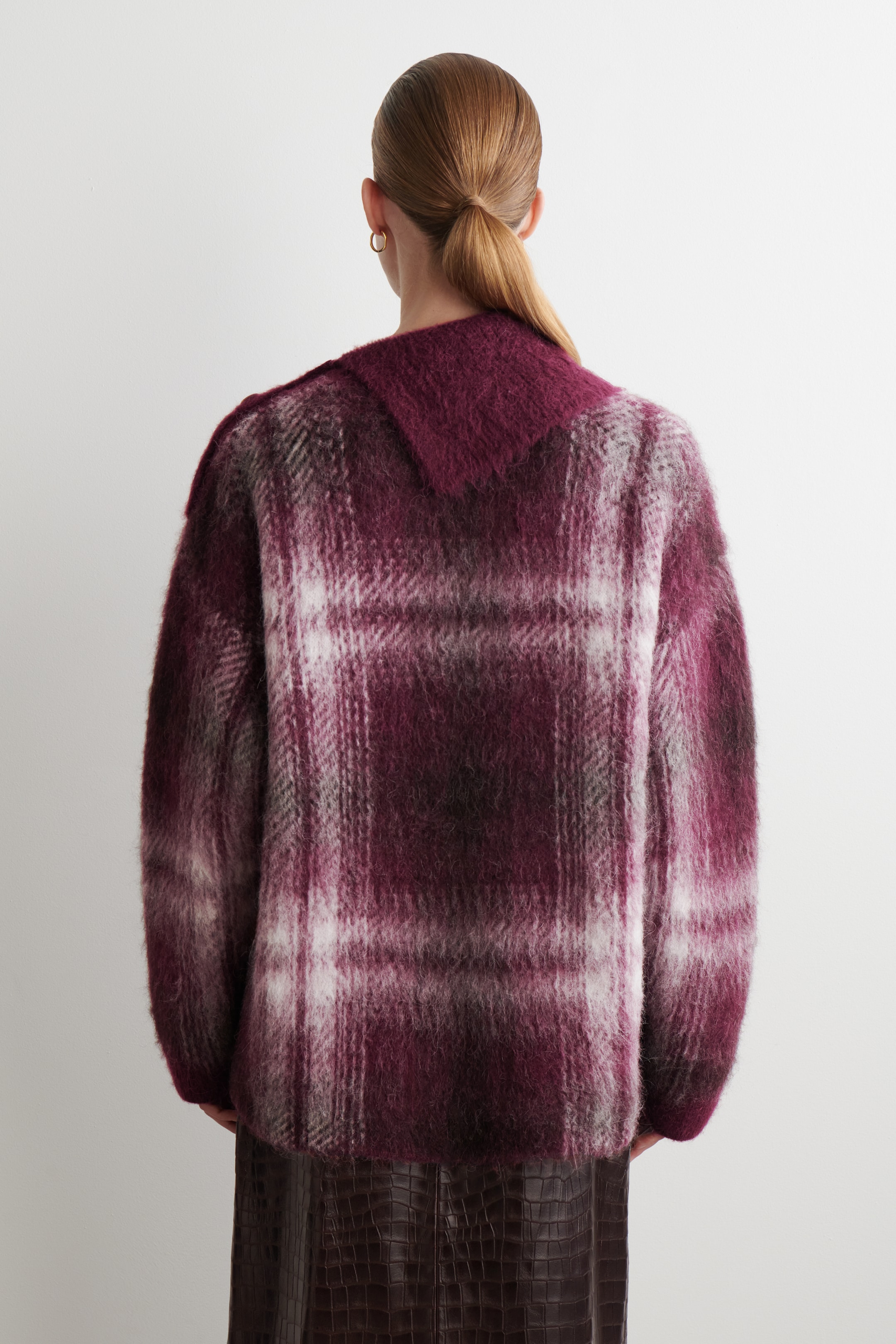Agrandir l'image: Un pull en plaid duveteux surdimensionné est montré de dos, avec un large col pointu se formant au niveau de l'encolure et un motif à carreaux dans des tons de rouge foncé, blanc et gris couvrant le matériau. Il est porté par-dessus une jupe texturée marron foncé.
