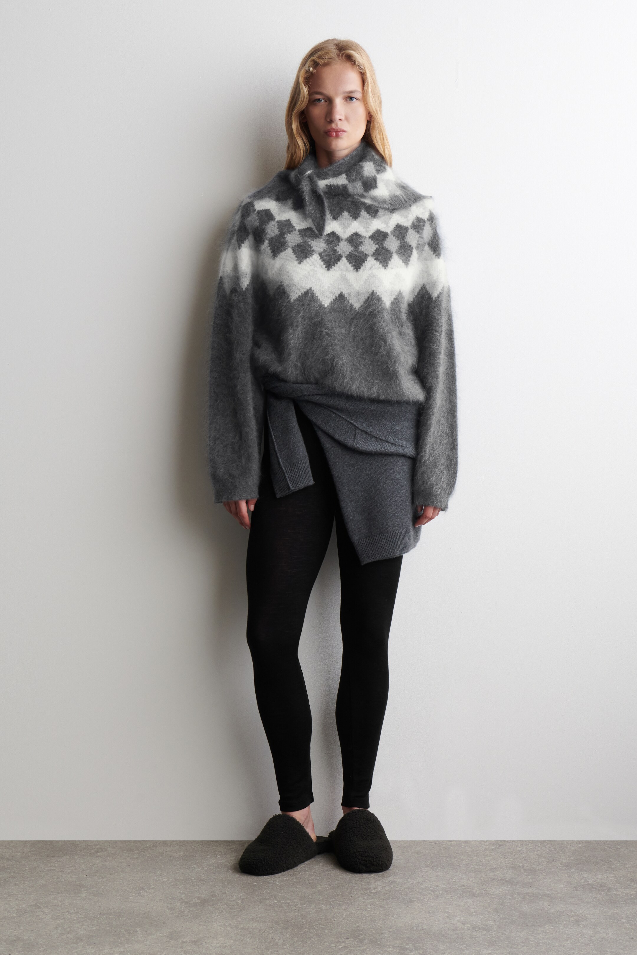 Ver imagen más grande: Un jersey Fair Isle de gran tamaño, de color gris y blanco borroso, presenta un cuello alto con cierre de botones. Una falda gris asimétrica está siendo usada sobre leggings negros. Se ven unas zapatillas de andar por casa de tono oscuro y texturizadas.