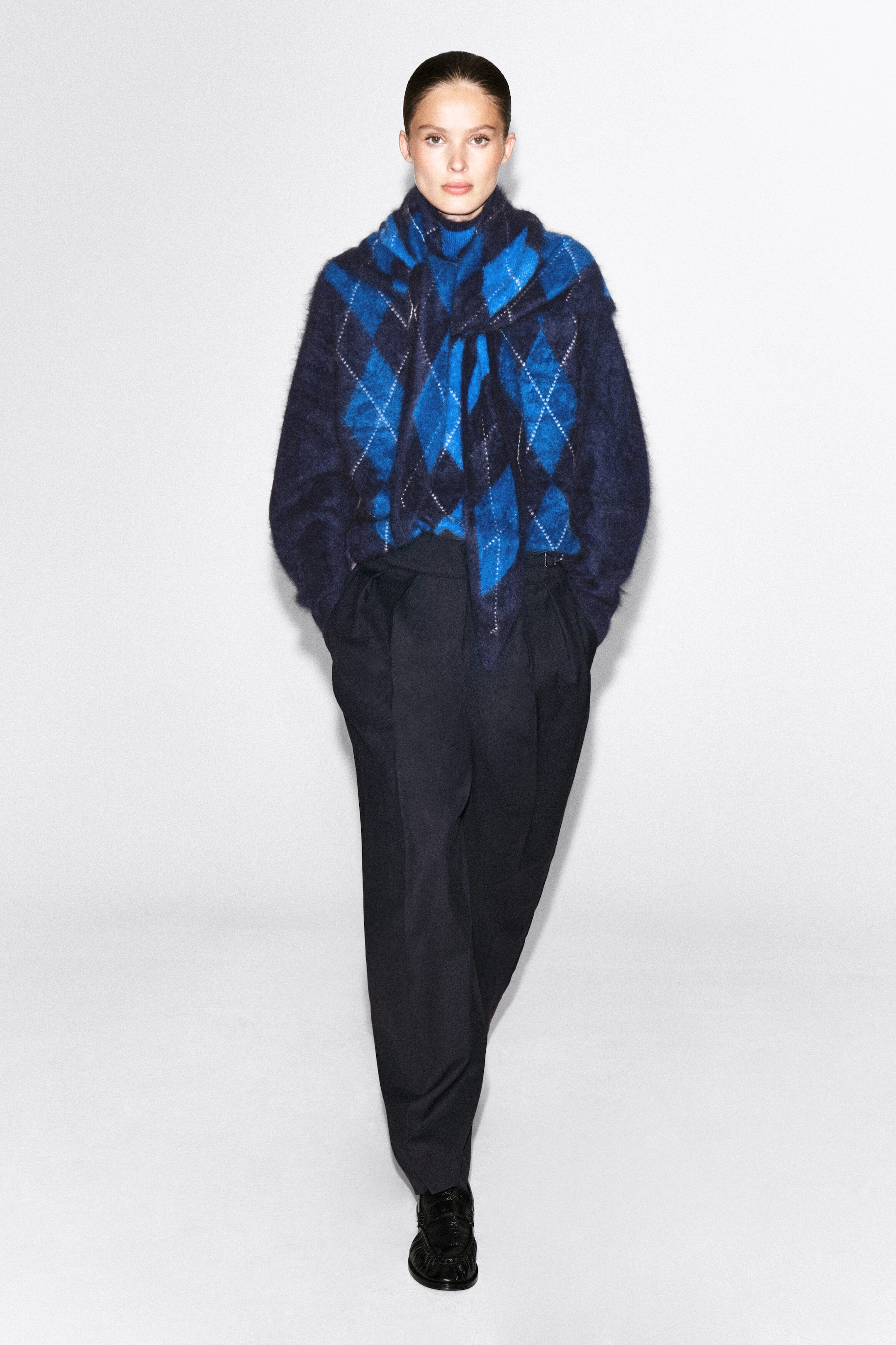 The model is wearing an argyle brushed cashmere funnel-neck jumper in shades of blue and navy. The jumper is styled with a matching argyle knit scarf tied around the shoulders and is tucked into dark, wide-leg trousers. The model is wearing an argyle brushed cashmere funnel-neck jumper in shades of blue and navy. The jumper is styled with a matching argyle knit scarf tied around the shoulders and is tucked into dark, wide-leg trousers.