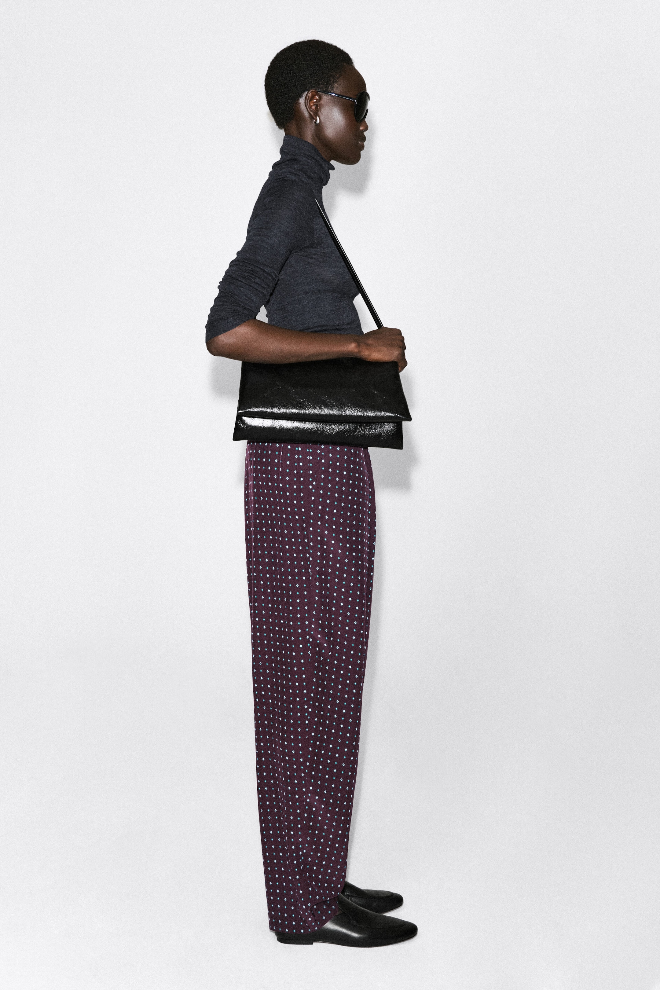 Ce pantalon droit à imprimé losanges, rehaussé d’un petit motif géométrique bleu clair sur une base prune profonde, tombe avec fluidité dans une silhouette décontractée tandis que le mannequin se tient de profil, tourné vers la droite. L’ourlet repose légèrement sur des chaussures plates noires, mettant en valeur sa longueur totale et sa structure souple.