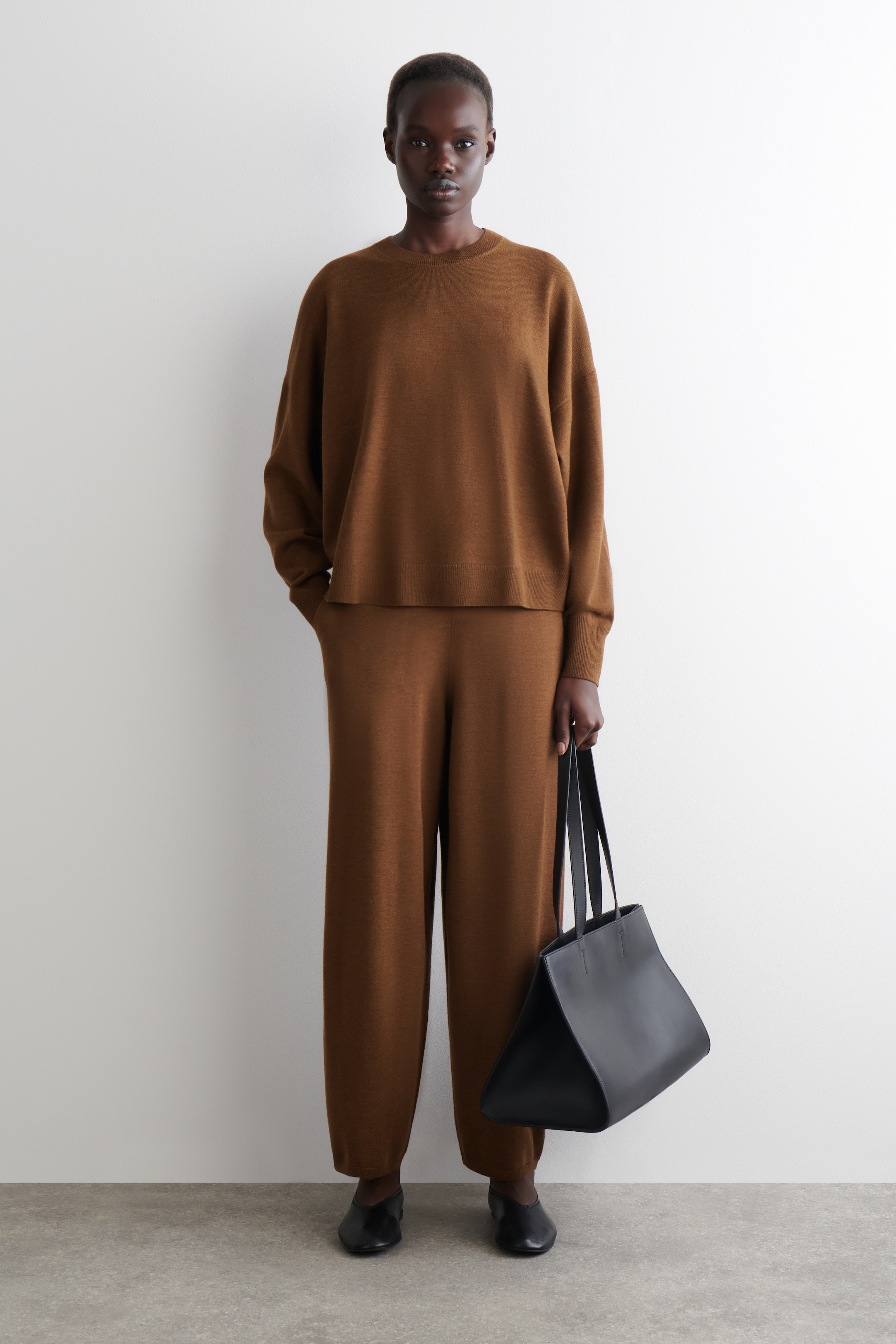 Agrandir l'image: Un ensemble en maille monochrome marron est porté, comprenant un pull à col rond à coupe décontractée avec des épaules tombantes et un pantalon large assorti. Un sac fourre-tout structuré noir est tenu, et des chaussures plates à bout pointu noires complètent la tenue.