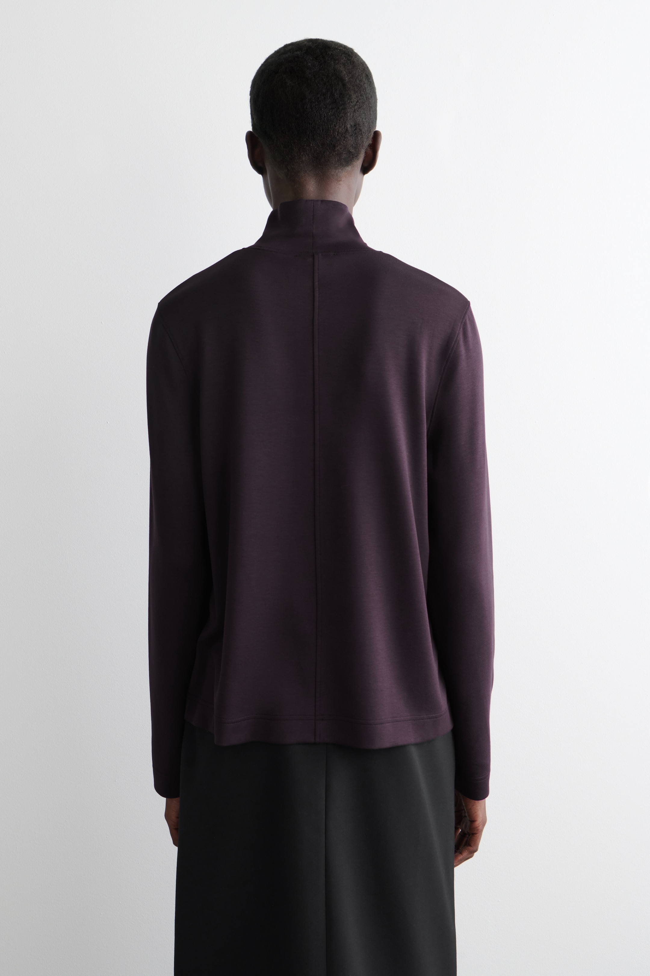 Agrandir l'image: Un haut à col montant violet foncé est présenté de dos, avec une couture centrale arrière proéminente et des manches longues. Le haut tombe avec un ourlet droit, superposé à une jupe noire.