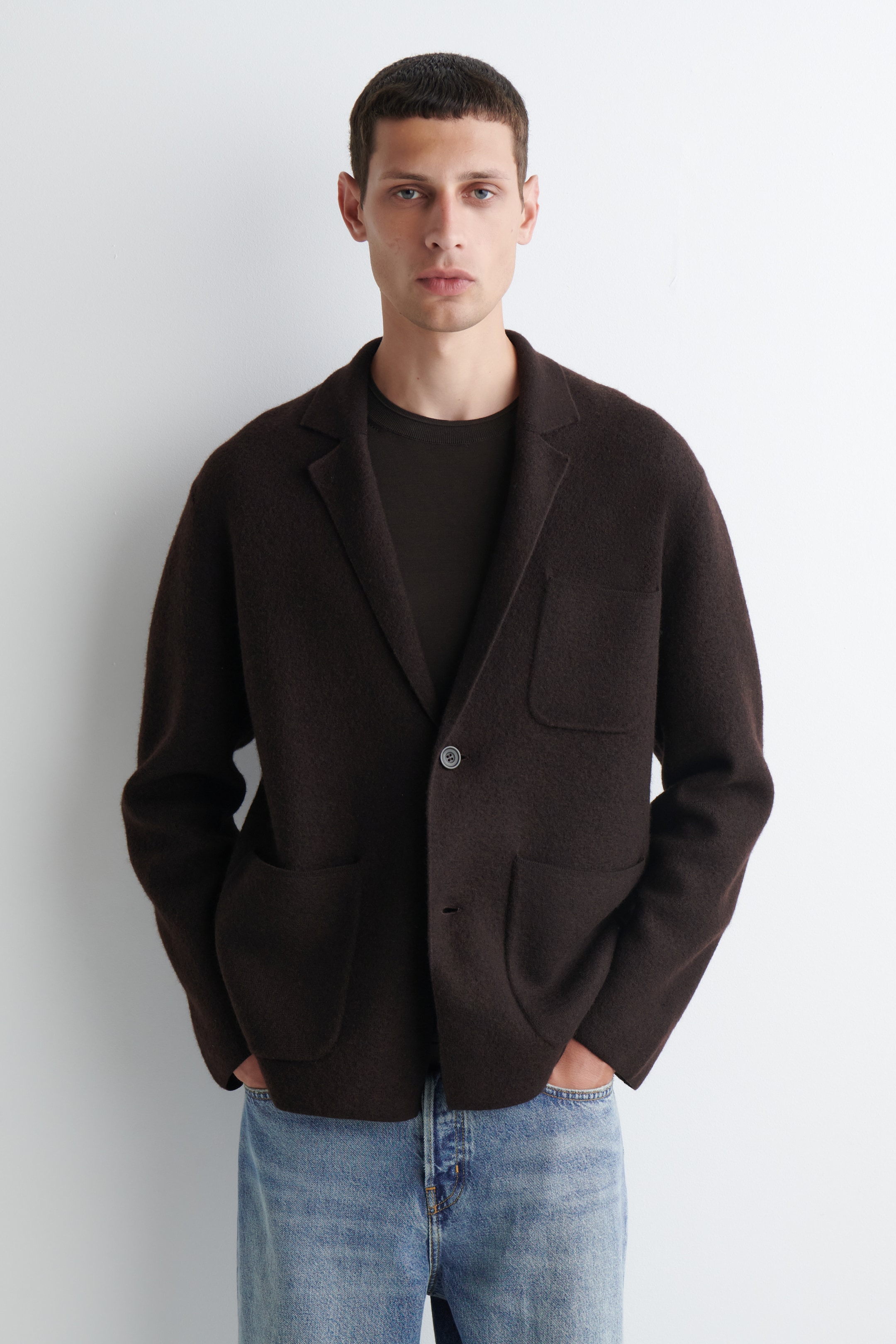 Größeres Bild ansehen: Ein dunkelbrauner einreihiger Blazer wird von vorne gezeigt, mit Kerbrevers, einem einzelnen dunklen Knopfverschluss, drei aufgesetzten Taschen, einer lockeren Passform und einer strukturierten, flauschigen Oberfläche, getragen über einem dunklen Rundhals-T-Shirt und hell gewaschenen Jeans.
