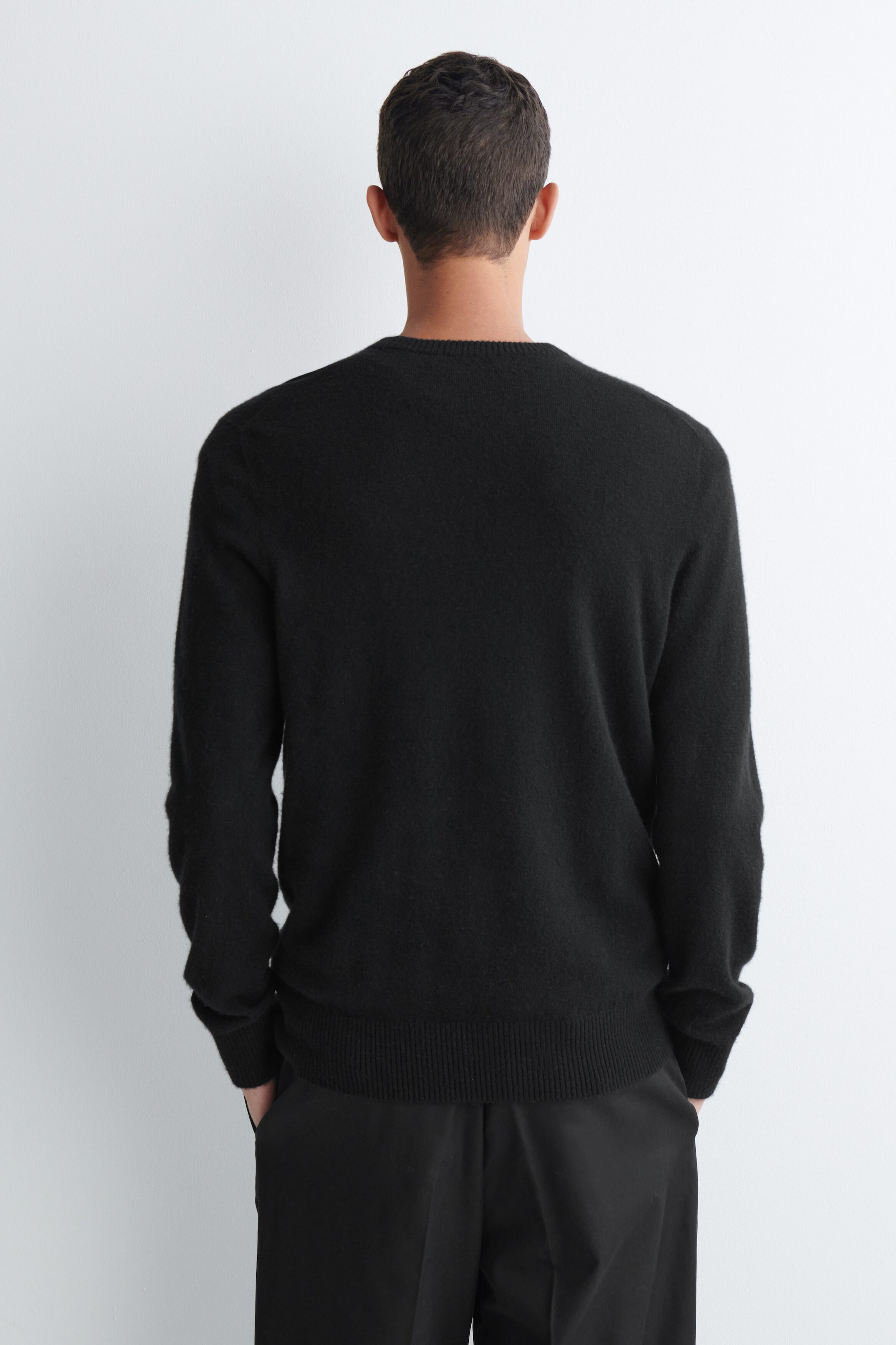 Ein schwarzer Pullover mit Rundhalsausschnitt und langen Ärmeln wird von dem Model getragen, das mit dem Rücken zur Kamera steht. Der Strickpullover verfügt über einen gerippten Ausschnitt, gerippte Bündchen und einen gerippten Saum und ist mit einer schwarzen Hose kombiniert. Ein schwarzer Pullover mit Rundhalsausschnitt und langen Ärmeln wird von dem Model getragen, das mit dem Rücken zur Kamera steht. Der Strickpullover verfügt über einen gerippten Ausschnitt, gerippte Bündchen und einen gerippten Saum und ist mit einer schwarzen Hose kombiniert.