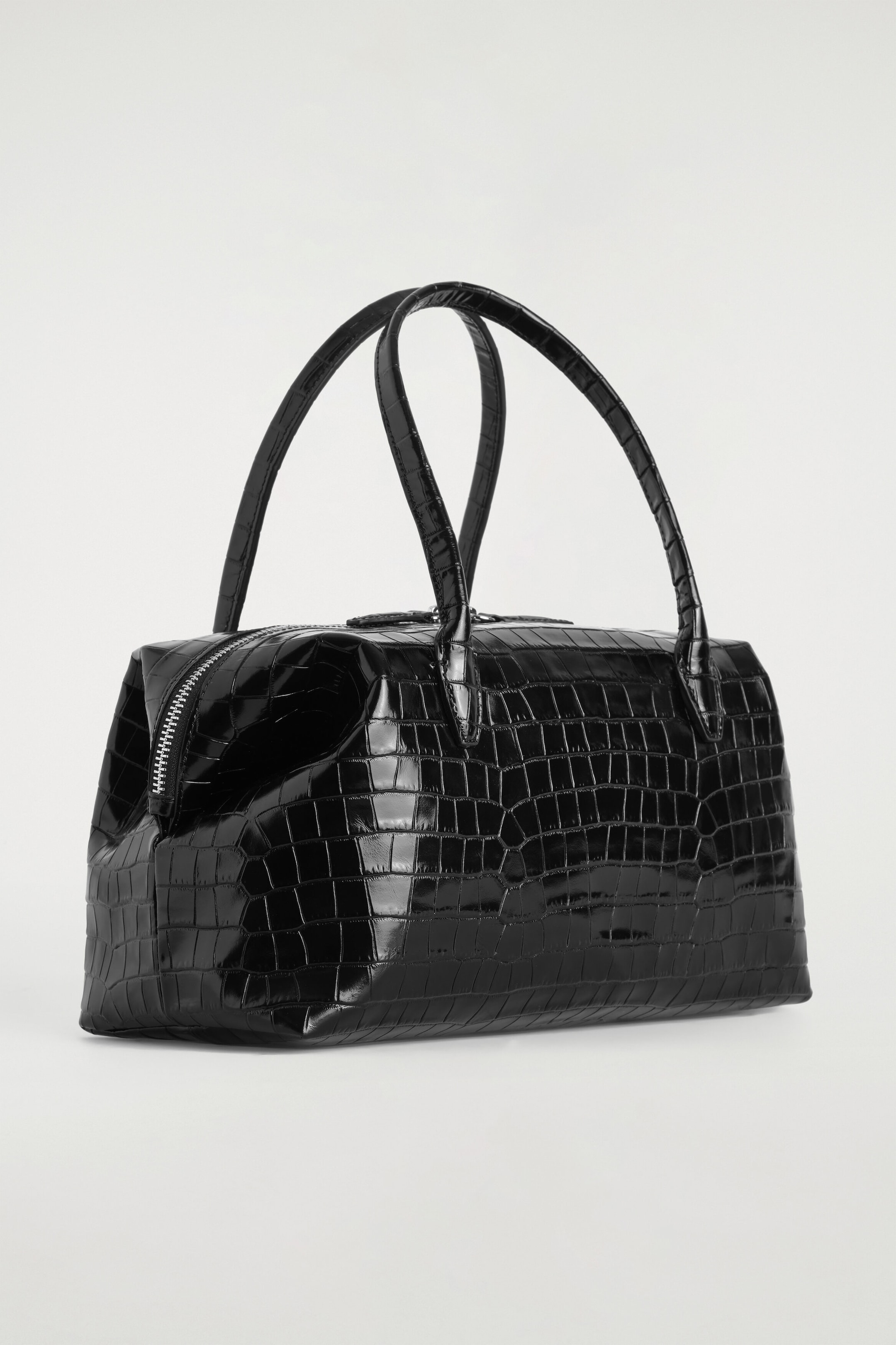 A black, glossy crocodile-embossed leather bowling bag is resting at a three-quarter angle, showcasing its sculptural shape with softly pinched sides. The bag is displaying two arched top handles and a silver-tone zip closure along the top. A black, glossy crocodile-embossed leather bowling bag is resting at a three-quarter angle, showcasing its sculptural shape with softly pinched sides. The bag is displaying two arched top handles and a silver-tone zip closure along the top.