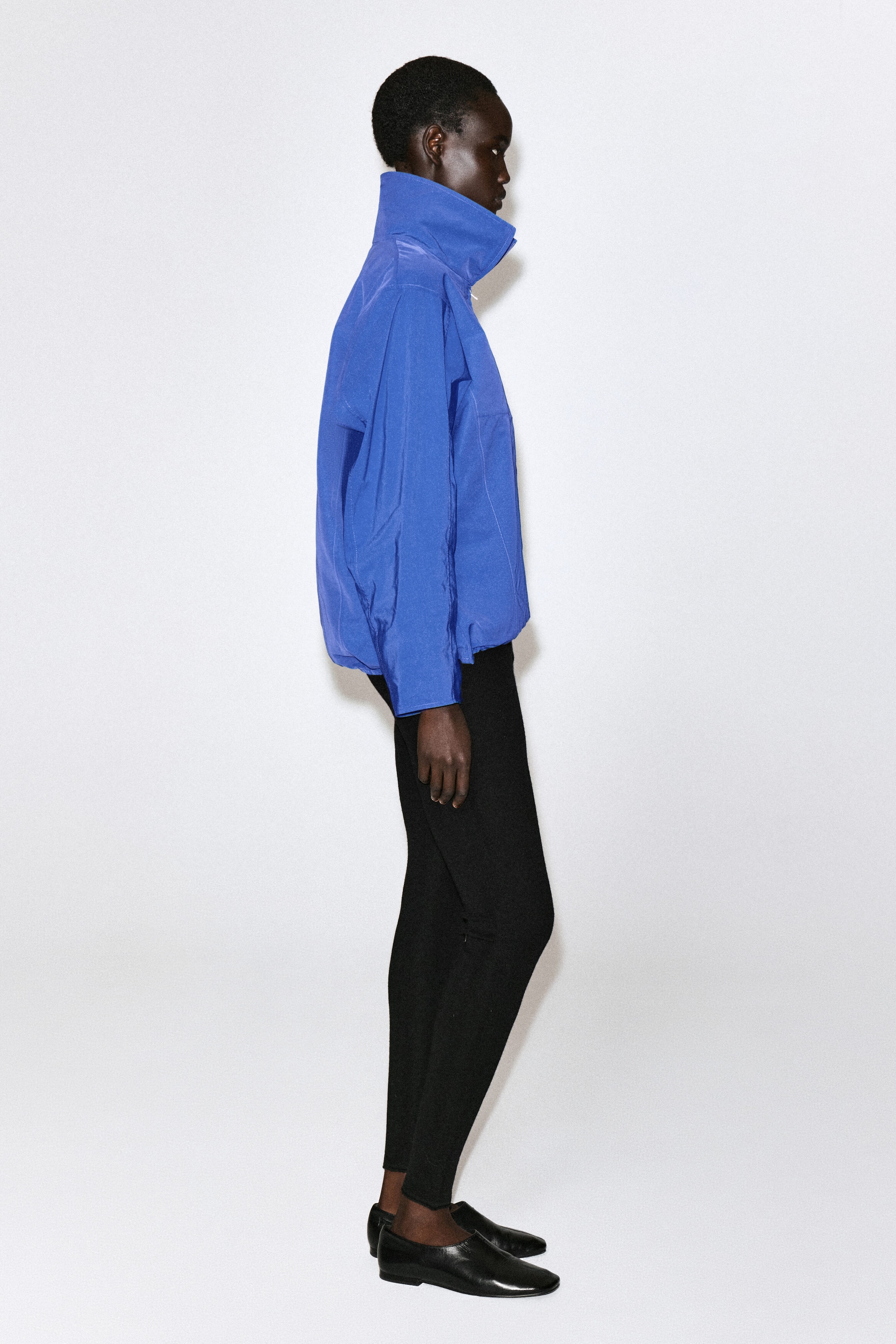 A royal blue zip-up jacket is shown in side profile facing right, highlighting its high funnel-neck collar, long sleeves, and elasticated drawstring hem. The jacket is layered over slim-fit black trousers and paired with black leather flat shoes. A royal blue zip-up jacket is shown in side profile facing right, highlighting its high funnel-neck collar, long sleeves, and elasticated drawstring hem. The jacket is layered over slim-fit black trousers and paired with black leather flat shoes.