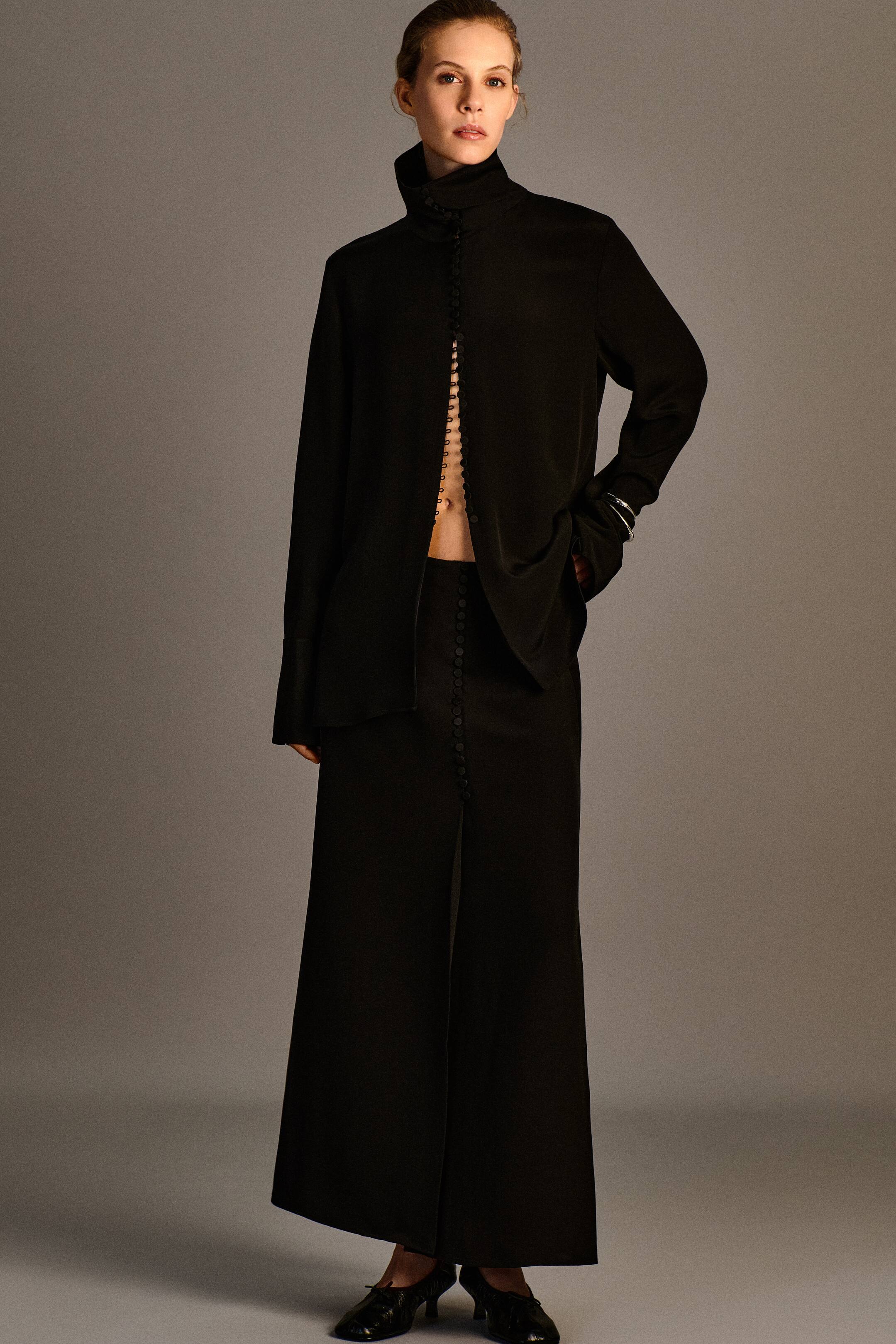 View larger image: The Button-Detail Silk-Crepe Shirt and Button-Detail Silk-Crepe Maxi Wrap Skirt are worn as a coordinated black set, shown from the front. The long-sleeved shirt features a high stand collar and a front button-and-loop fastening left partially open at the waist, while the matching high-waisted maxi skirt showcases a vertical row of fabric-covered buttons along its front wrap fastening.