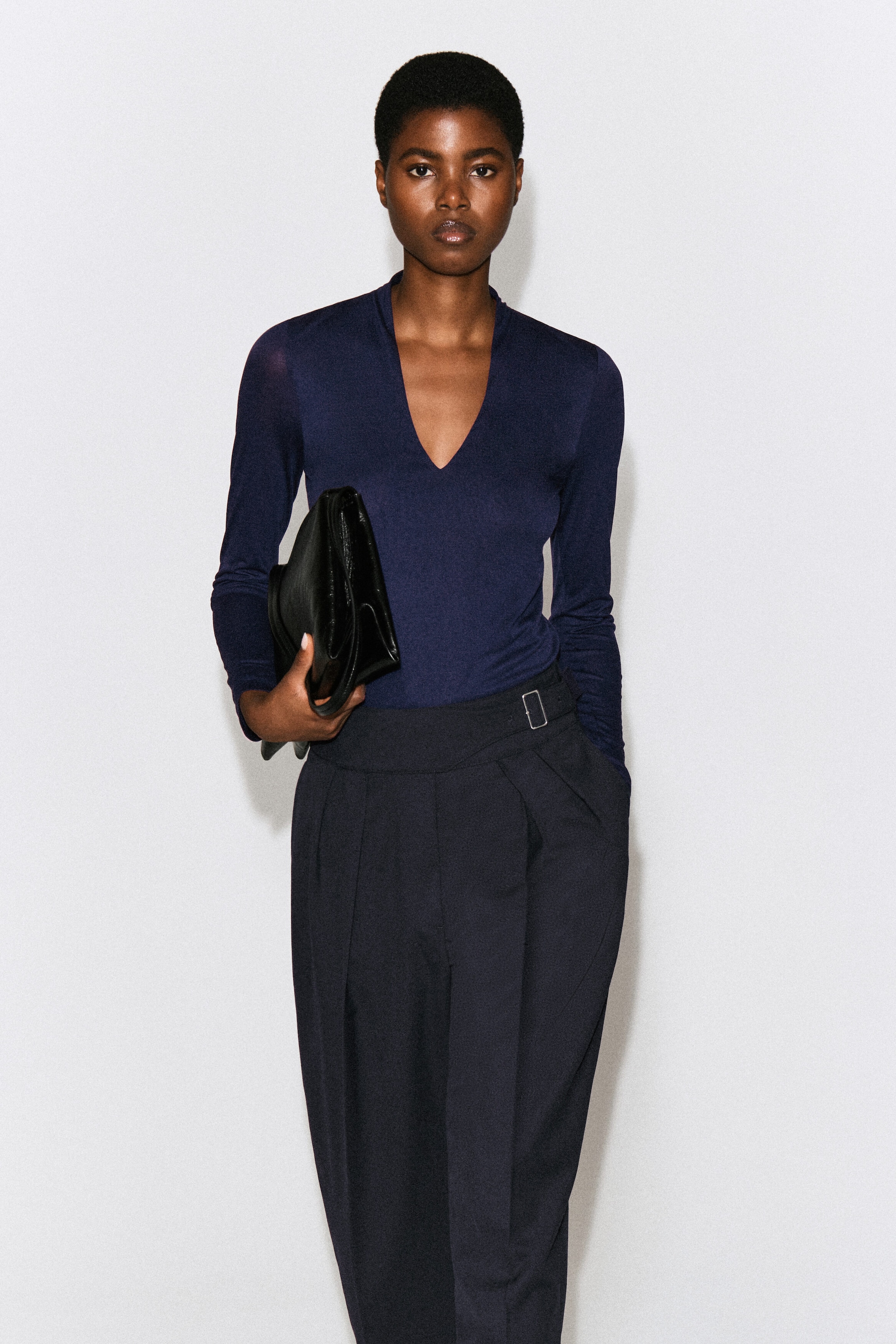 The model is wearing a dark navy blue long-sleeved V-neck top tucked into dark pleated trousers. The model is holding a black leather clutch under one arm. The model is wearing a dark navy blue long-sleeved V-neck top tucked into dark pleated trousers. The model is holding a black leather clutch under one arm.
