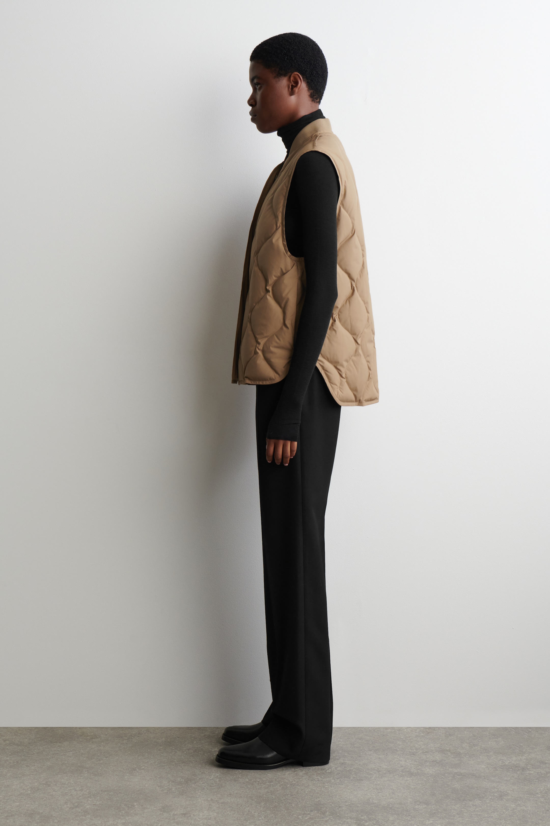 View larger image: A tan quilted vest is being displayed from a side profile, featuring a wavy diamond pattern and a rounded hem. It is being layered over a black turtleneck and black wide-leg trousers, showcasing its hip-length fit.