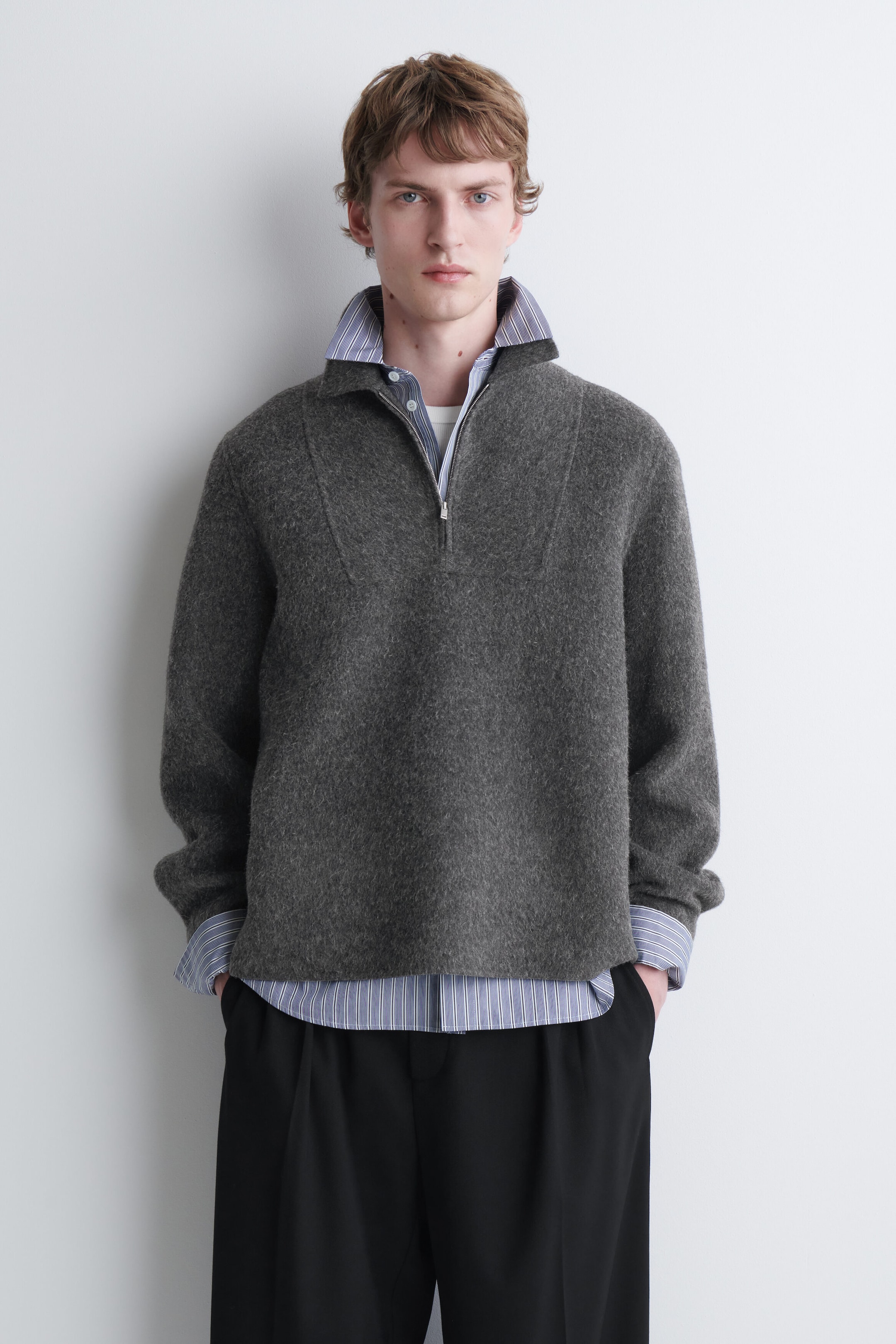 A gray textured wool half-zip sweater is being worn by the model, layered over a blue and white striped collared shirt and black trousers. The sweater features a high collar with a metal zipper that is left slightly open to reveal the layered shirts underneath.