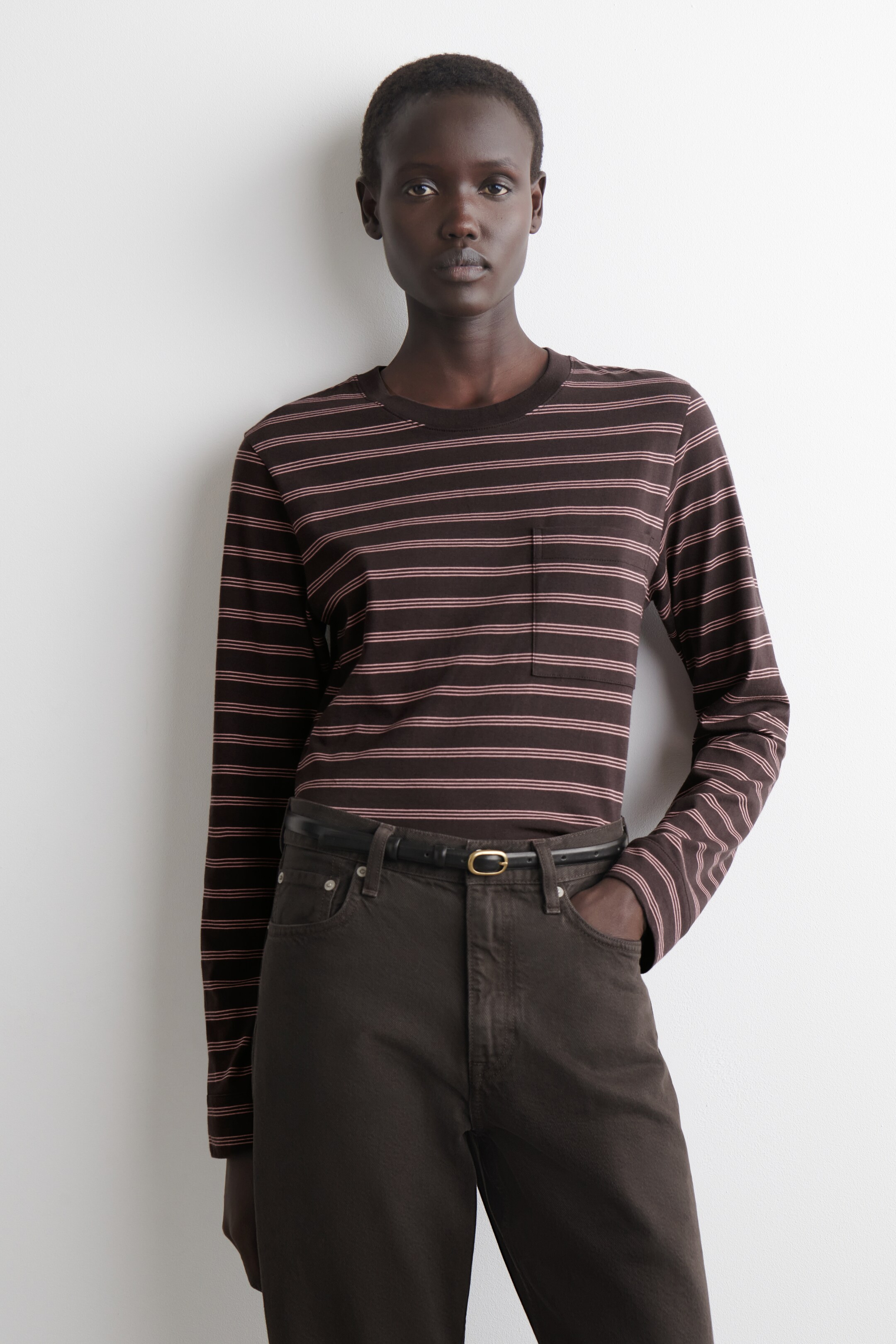 Zobacz większy obraz: Ciemnobrązowa koszulka z długim rękawem w cienkie różowe poziome paski, noszona z okrągłym dekoltem i kieszonką na lewej piersi. Włożona w ciemnobrązowe spodnie, zabezpieczone cienkim ciemnym paskiem z owalną złotą klamrą, wszystko skierowane do przodu.