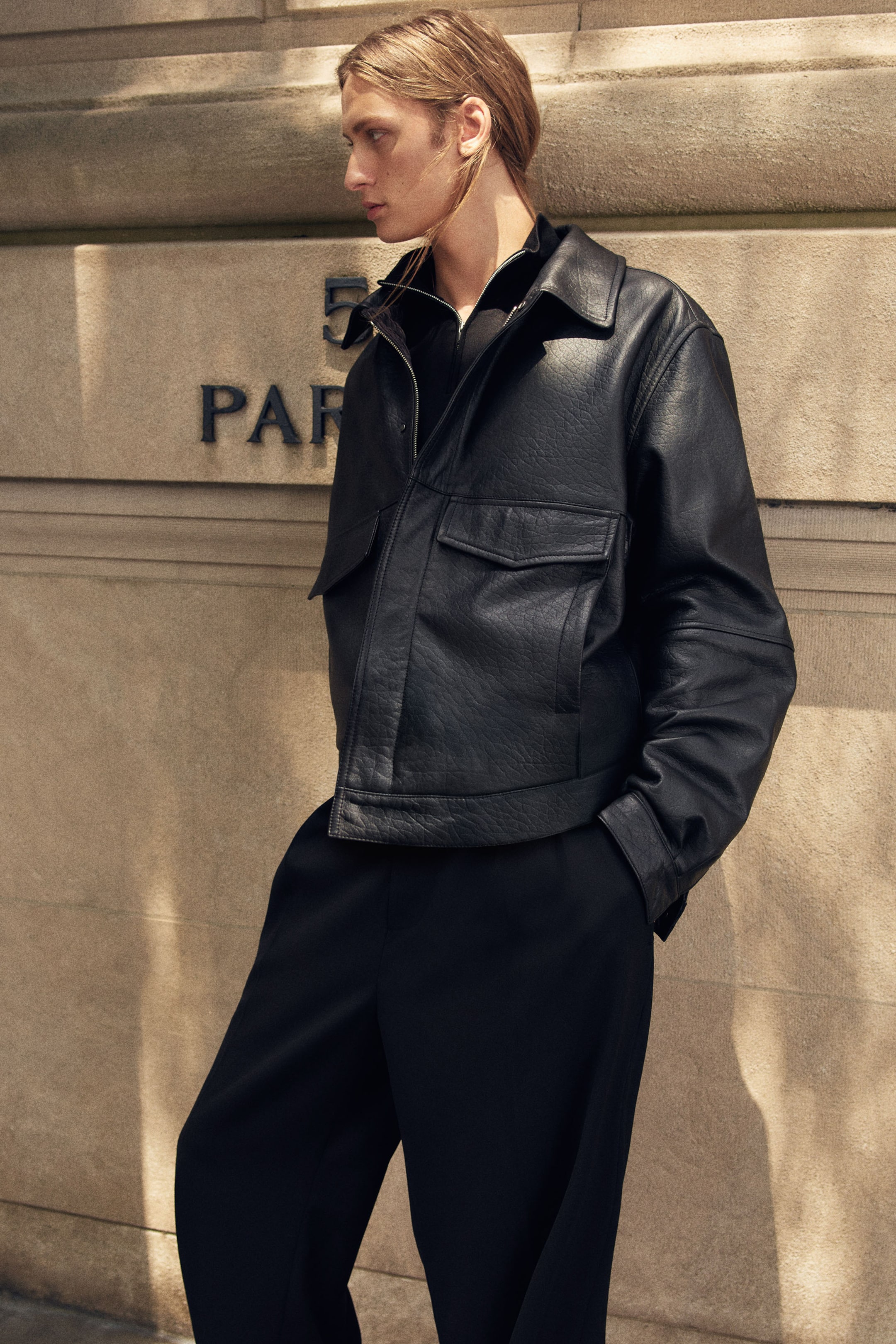 View larger image: A black leather flight jacket with a textured grain finish is shown from a three-quarter angle, highlighting its relaxed fit and structured design. The jacket features a classic spread collar, a front zip closure styled slightly open over a black zip-up collar, and prominent chest pockets with flap closures.