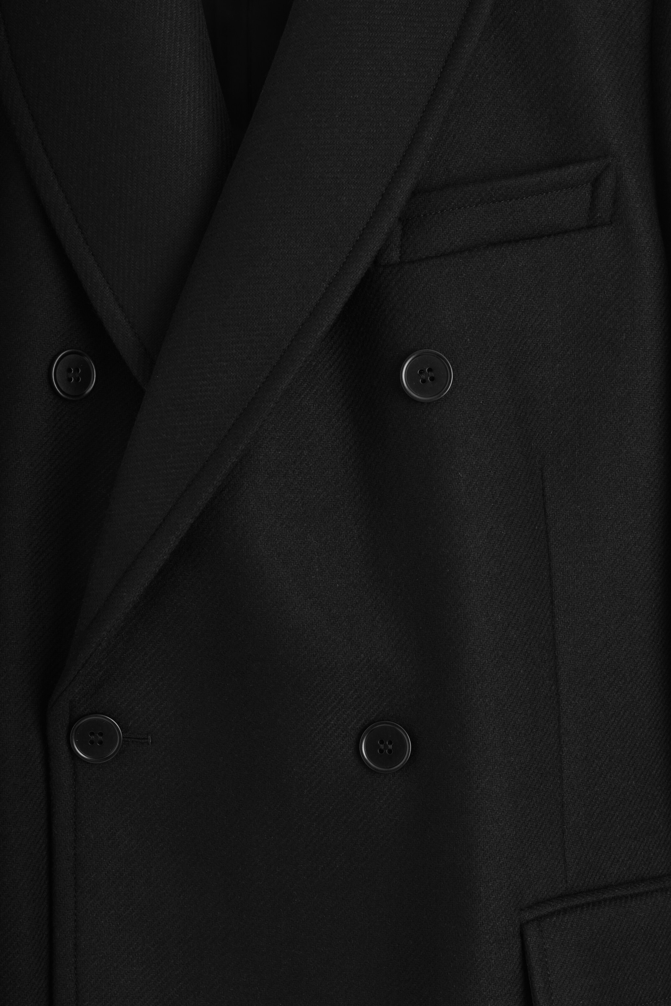 Visualizza immagine più grande: Un cappotto nero doppiopetto viene presentato frontalmente, mostrando bottoni scuri, revers a dente, un tessuto con una texture sottile e una tasca a filetto singola sul lato destro.