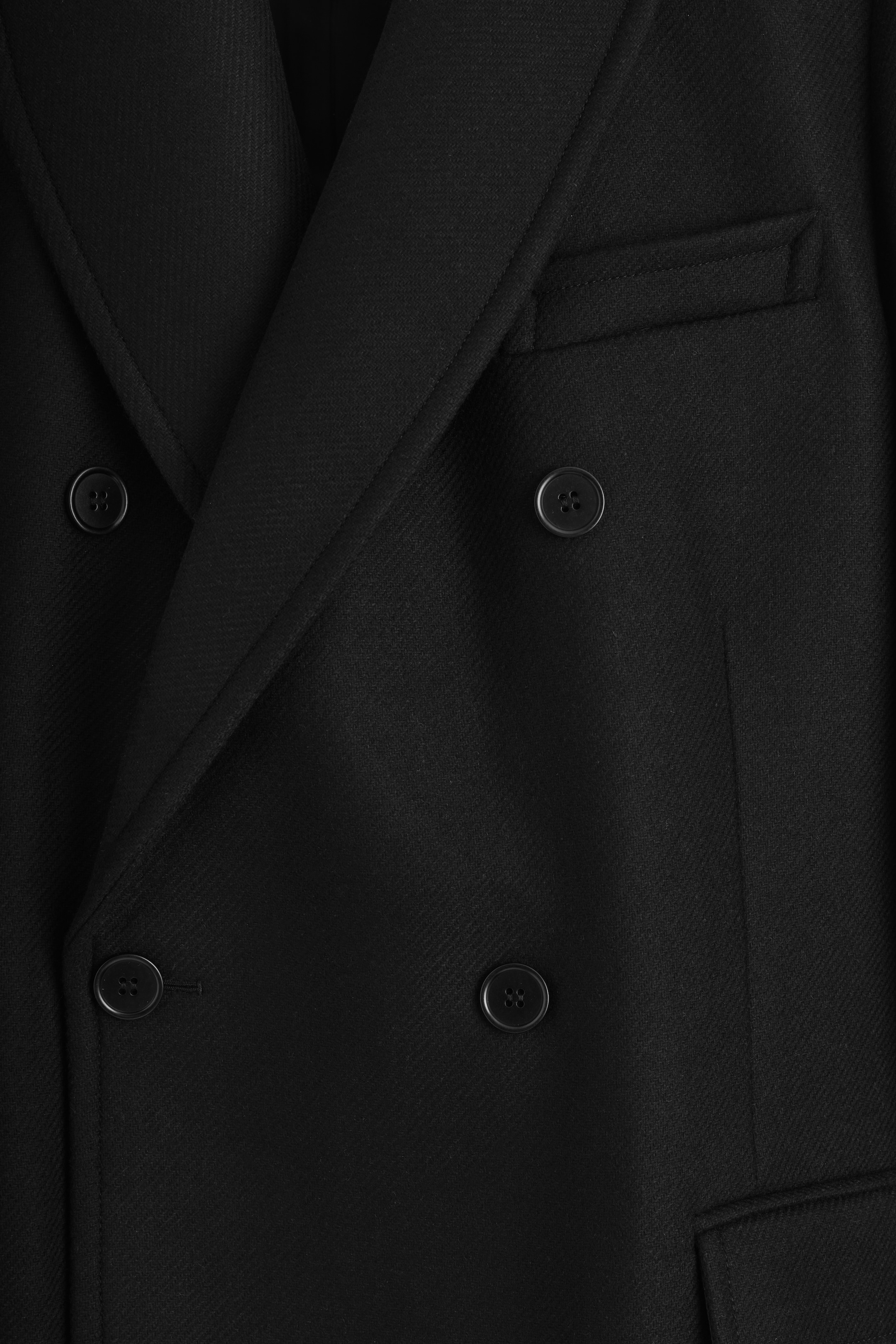 Visualizza immagine più grande: Un cappotto nero doppiopetto viene presentato frontalmente, mostrando bottoni scuri, revers a dente, un tessuto con una texture sottile e una tasca a filetto singola sul lato destro.