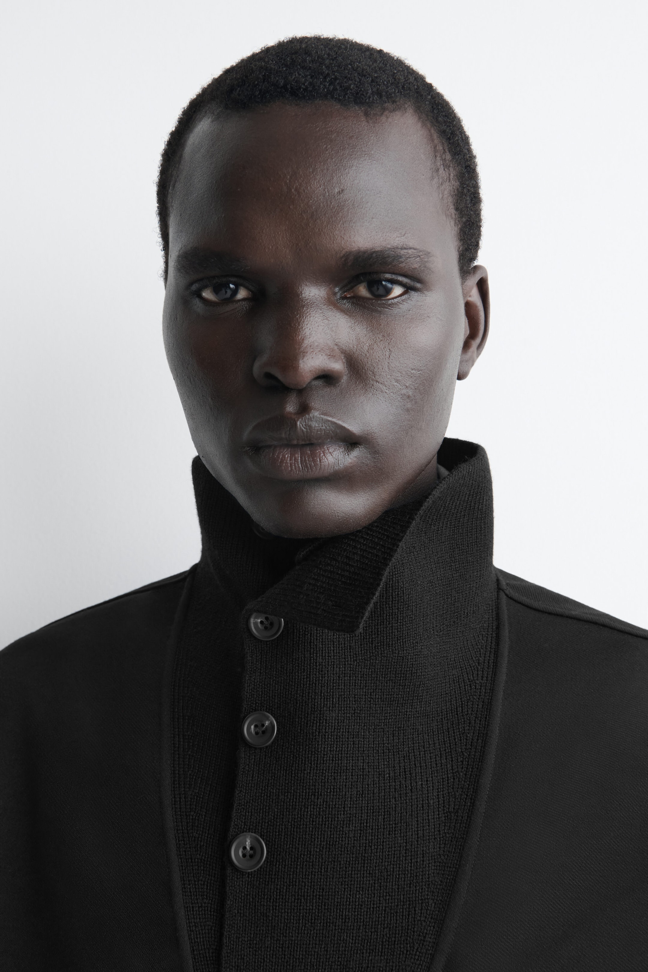 A black jacket features a high ribbed-knit funnel neck collar and a front button closure with three dark buttons. The garment showcases a contrasting smooth, structured fabric on the shoulders and body, presented in a close-up, front-facing view.