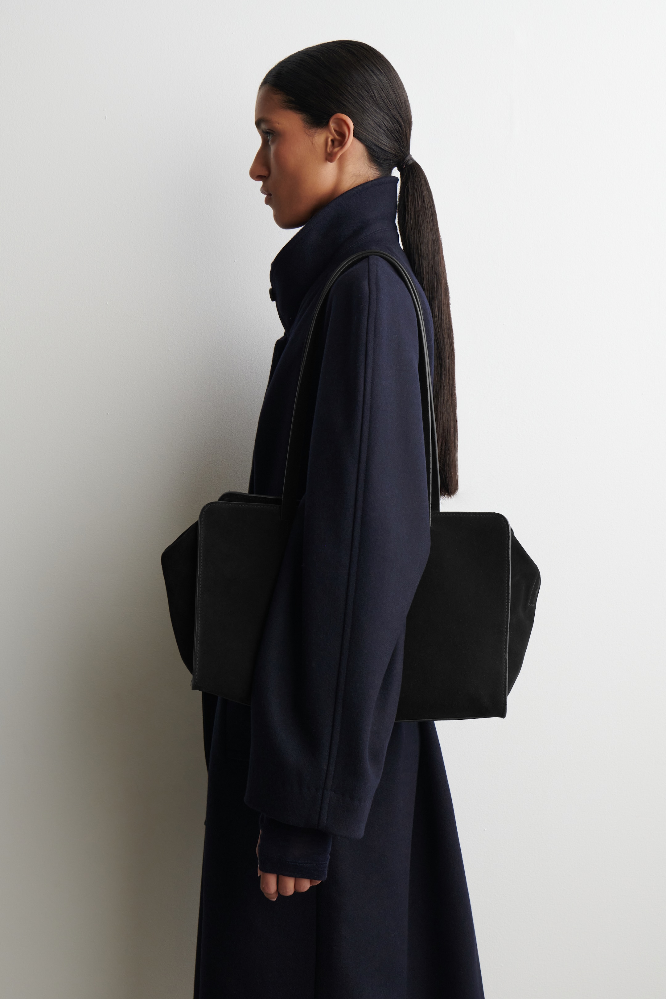 Visualizza immagine più grande: Un cappotto blu scuro viene indossato di profilo, caratterizzato da una silhouette dritta con un colletto alto e maniche lunghe e dritte. Una borsa rettangolare nera è appesa a una tracolla sopra il cappotto.