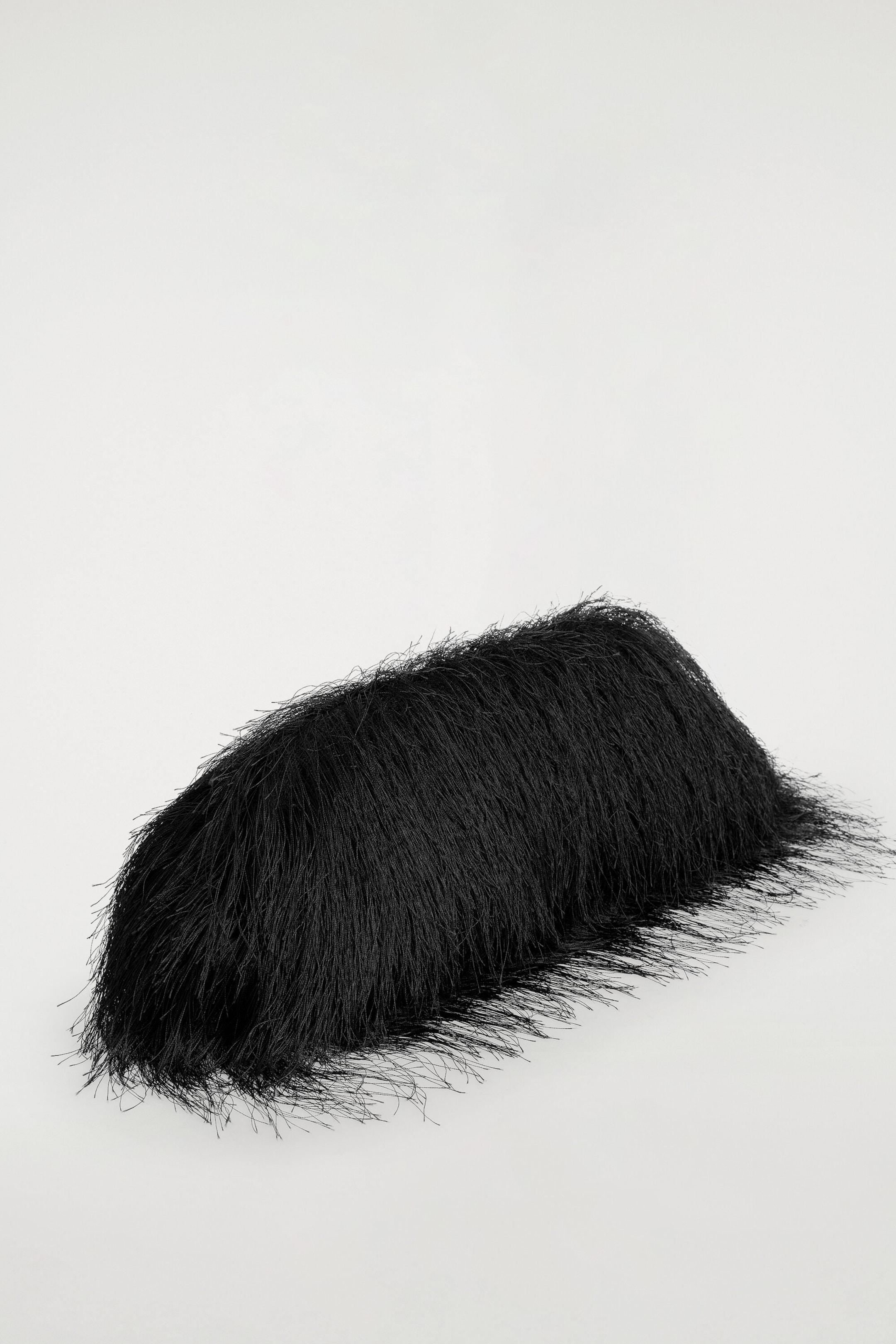 A black fringed clutch bag is shown from a three-quarter front angle, highlighting its elongated, rounded shape. The entire surface is covered in long, fine black fringe, creating a densely textured, shaggy appearance.