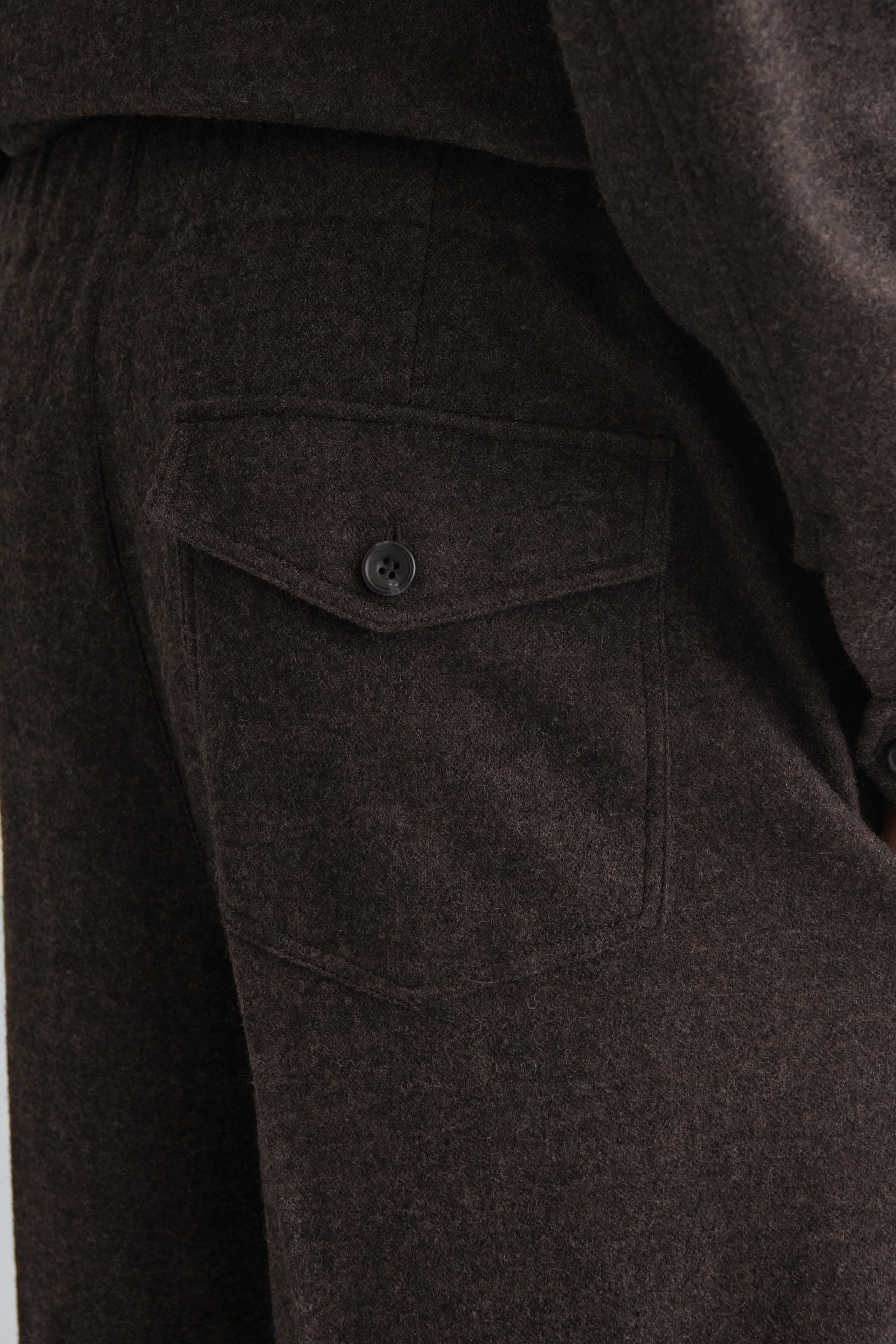Visualizza immagine più grande: Pantaloni in misto lana marrone scuro vengono presentati da una vista posteriore leggermente angolata, con il loro tessuto strutturato chiaramente visibile. Una tasca a toppa con chiusura a patta è evidenziata sul retro, e un bottone scuro fissa la patta.