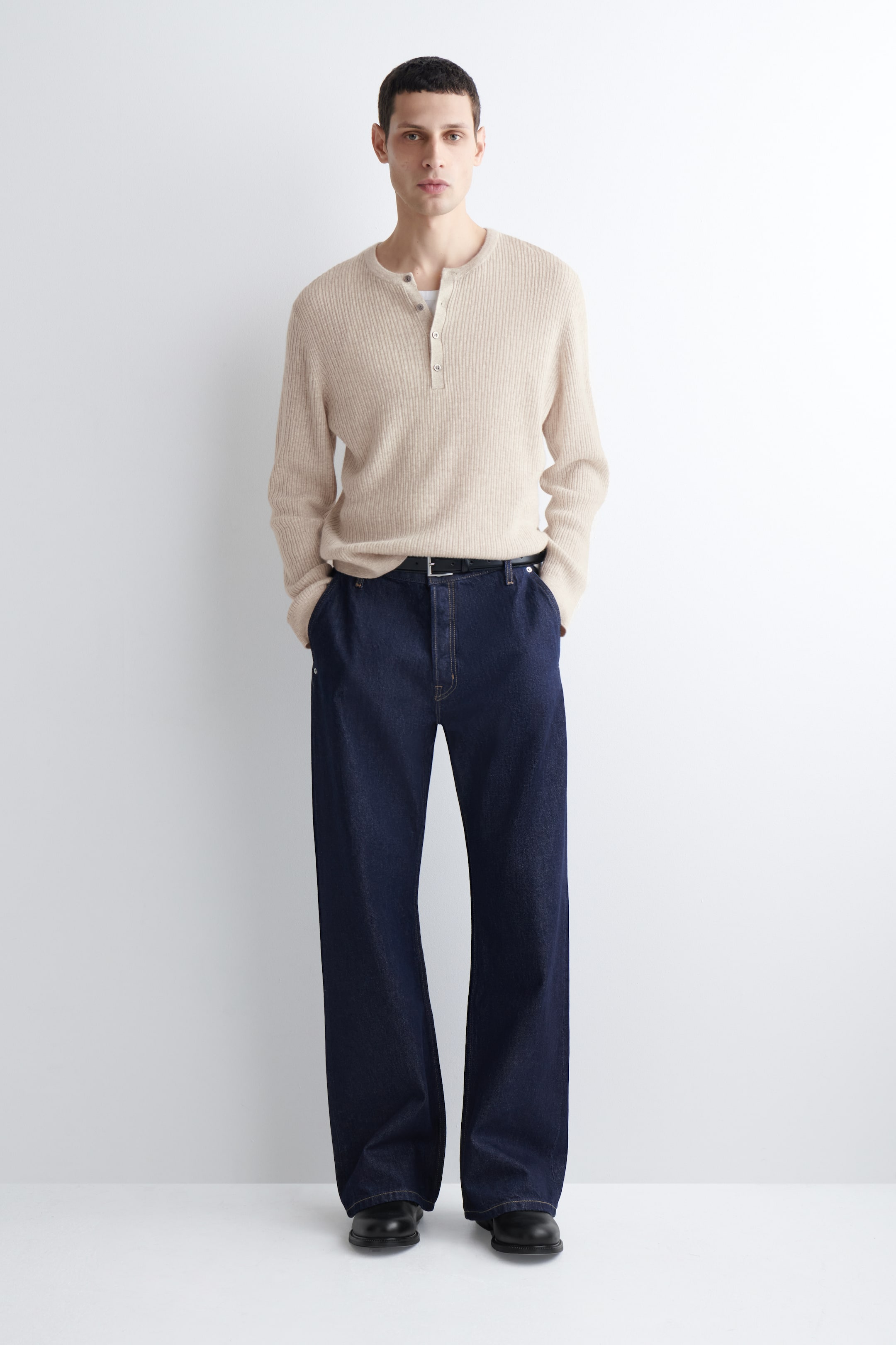 Grotere afbeelding bekijken: Een beige geribde Henley trui wordt gedragen over een lichtere onderhemd, met een drieknoopsluiting en lange mouwen. Het wordt gecombineerd met donkerblauwe wijde jeans, die in de taille worden vastgemaakt met een zwarte riem, en zwarte schoenen zijn zichtbaar.